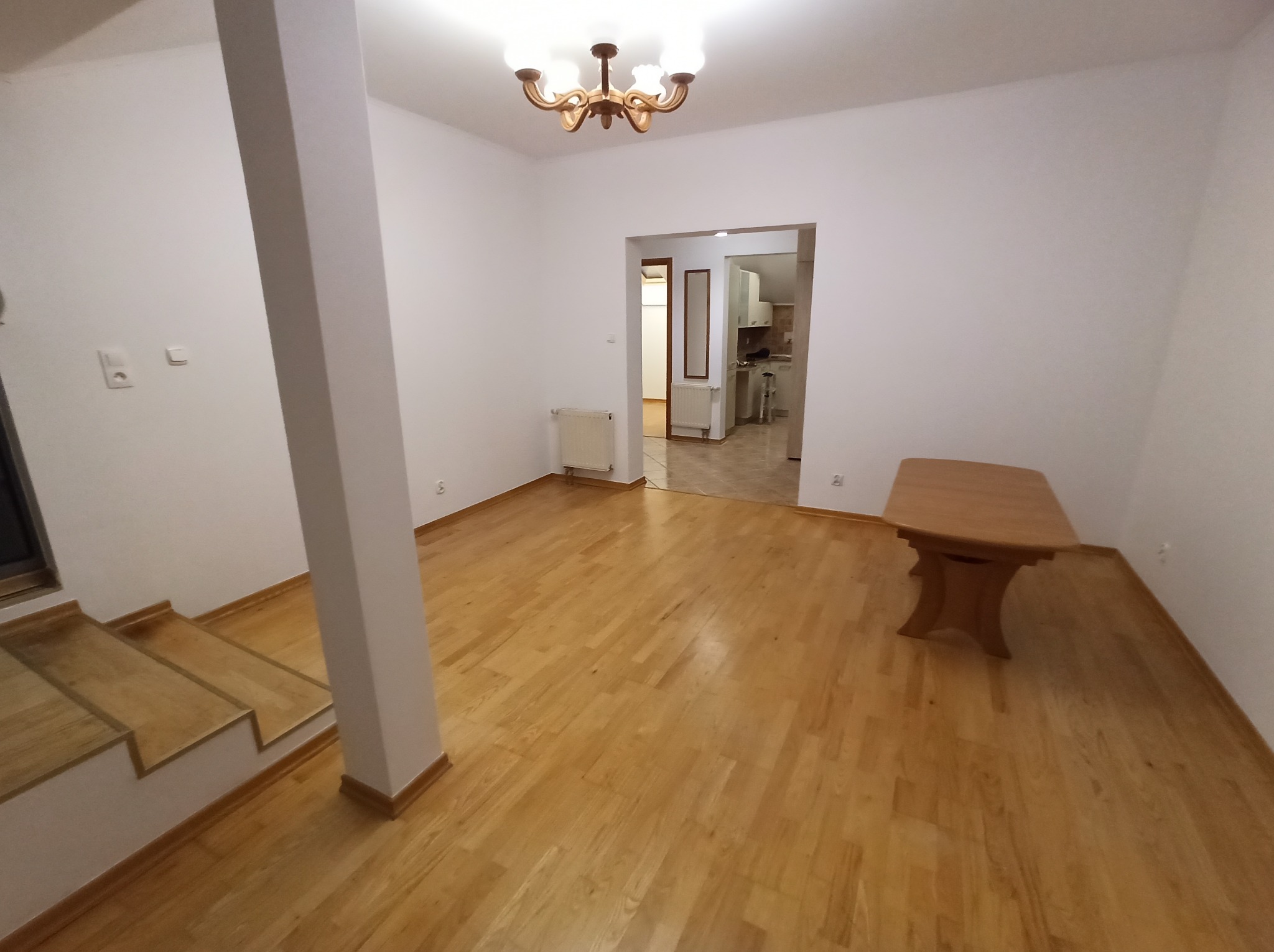 Puste, jasne wnętrze po malowaniu: białe ściany, parkiet z drewna, widoczny fragment schodów i kolumna. W tle widok na kuchnię, klasyczny żyrandol.