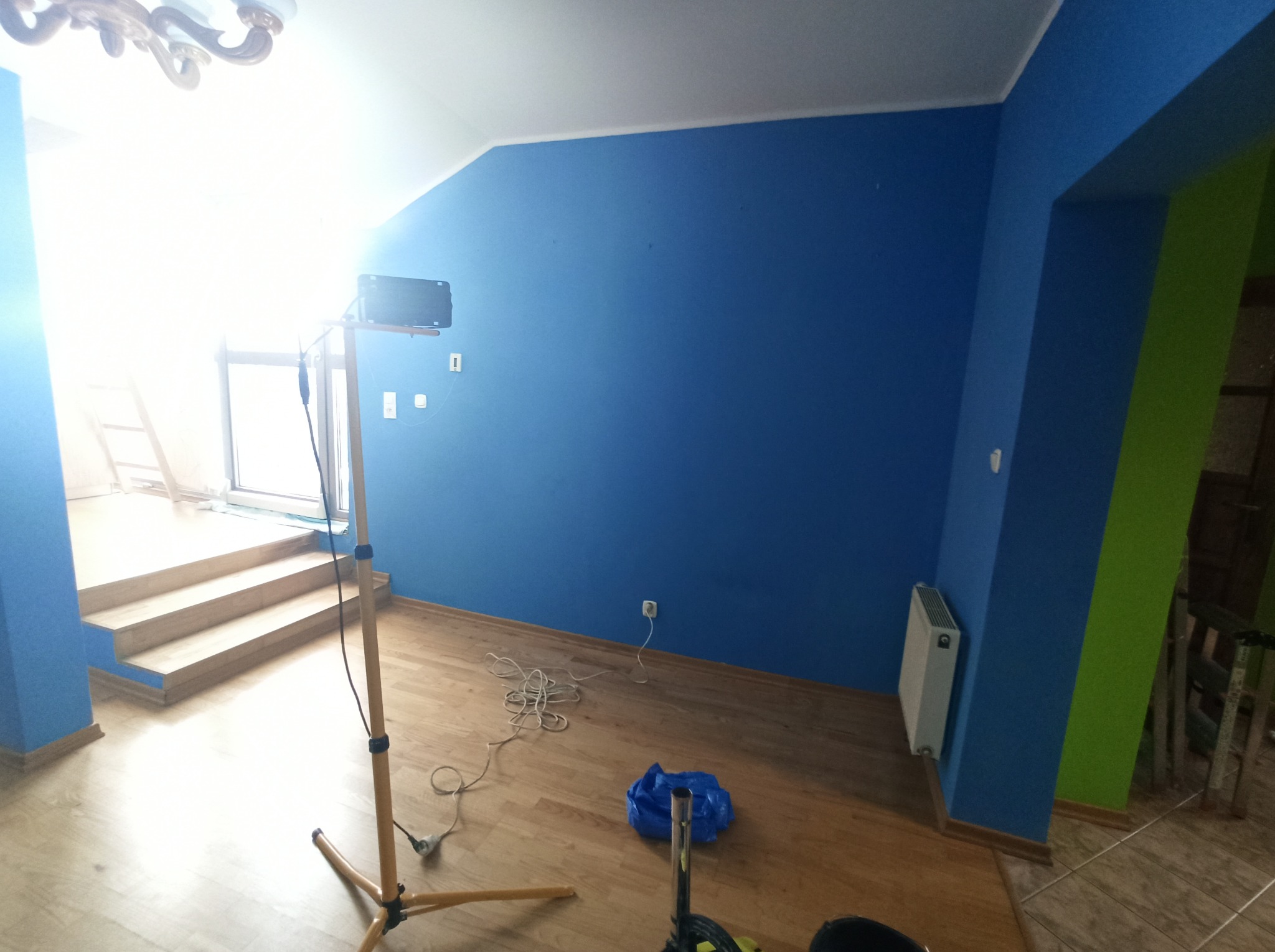 Pokój po malowaniu ścian na niebiesko i zielono, widoczny statyw z lampą oświetlającą. Podłoga drewniana, grzejnik przy ścianie. Częściowo widoczne schody.