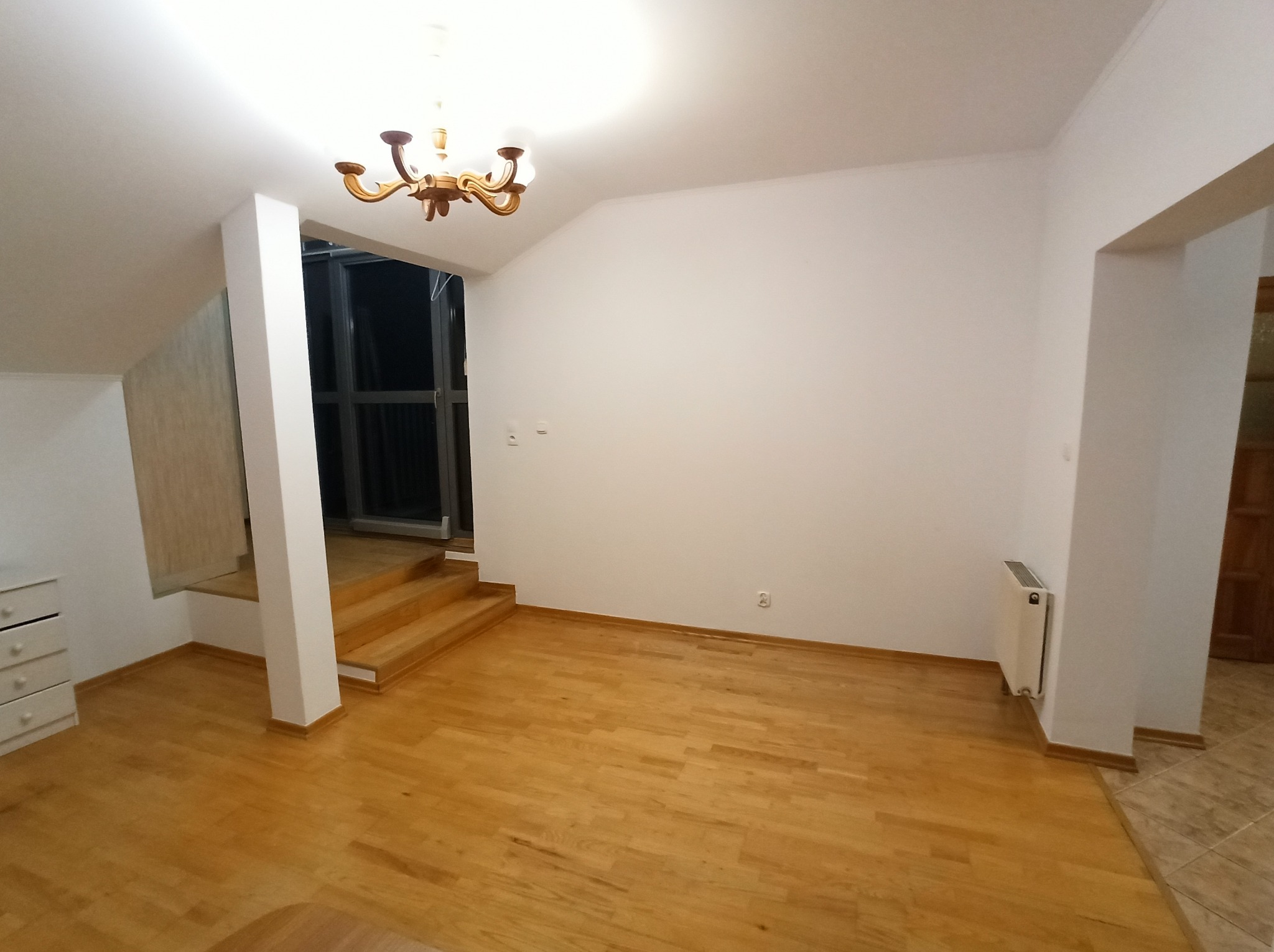 Jasne, przestronne wnętrze po malowaniu: białe ściany, drewniana podłoga, schody i dekoracyjny żyrandol. Widoczny grzejnik i gniazdko elektryczne.
