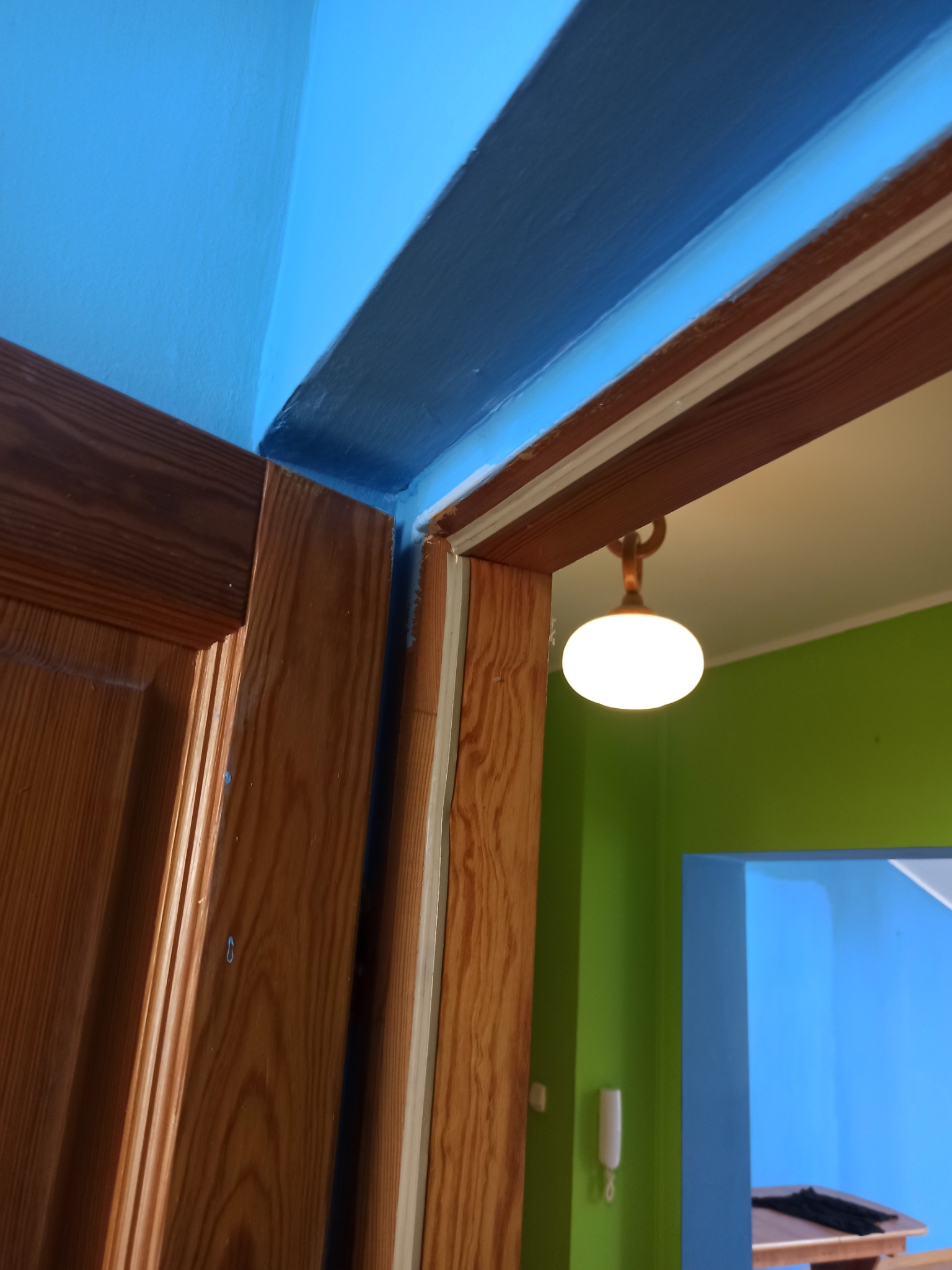 Fragment odnowionego mieszkania: świeżo pomalowany na niebiesko sufit i ściana przy drewnianych drzwiach, widok na pokój z zieloną ścianą i lampą.