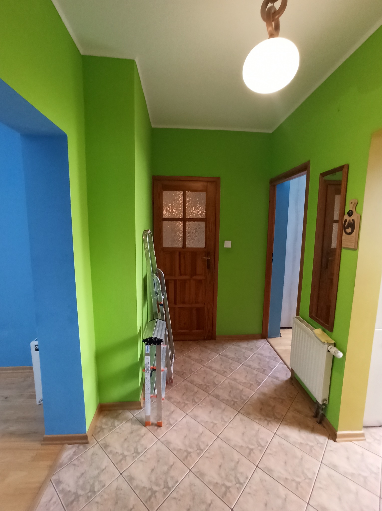 Wnętrze po malowaniu: żywe, zielone ściany, błękitne wnęki i drewniane drzwi. Aluminiowa drabina oparta o ścianę. Podłoga wyłożona płytkami w odcieniach beżu.