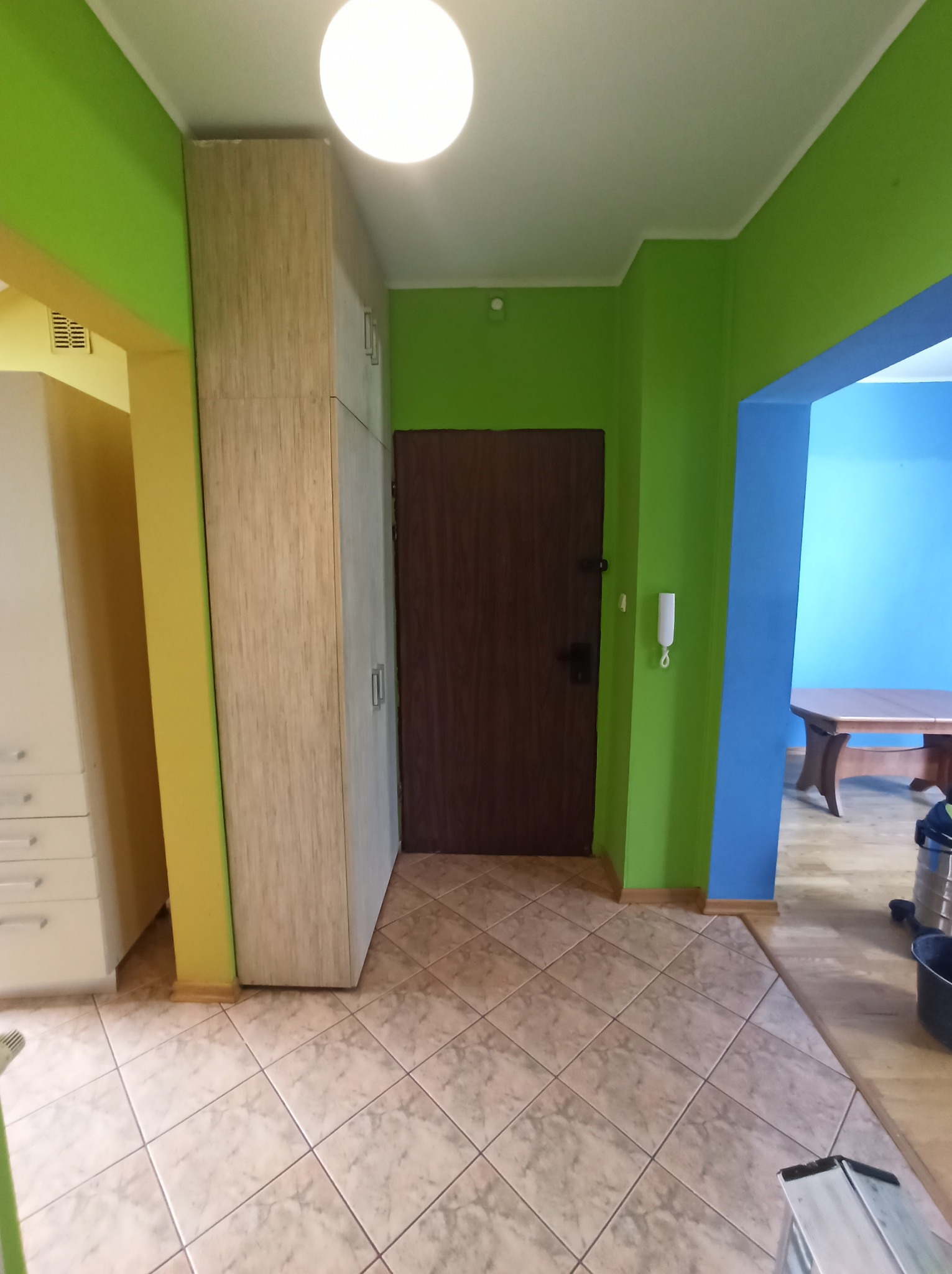 Wnętrze mieszkania po malowaniu: ściany w odcieniach zieleni i błękitu, brązowe drzwi, beżowa podłoga z płytek, białe szafy. Widok na pomalowane pomieszczenie.