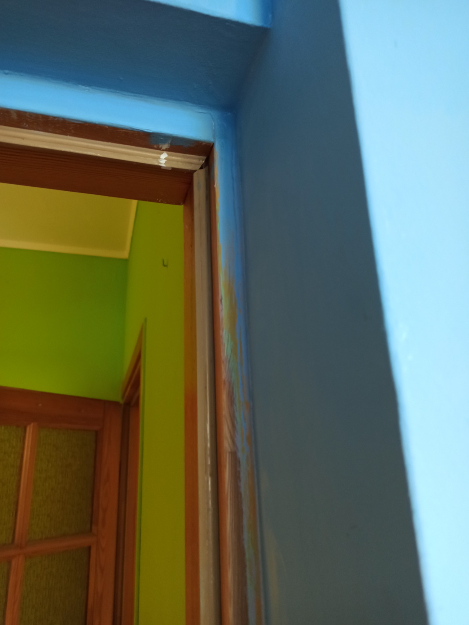 Fragment futryny drzwiowej w trakcie malowania na niebiesko, z widocznymi śladami farby i drewnianą ramą. W tle zielona ściana i drzwi.