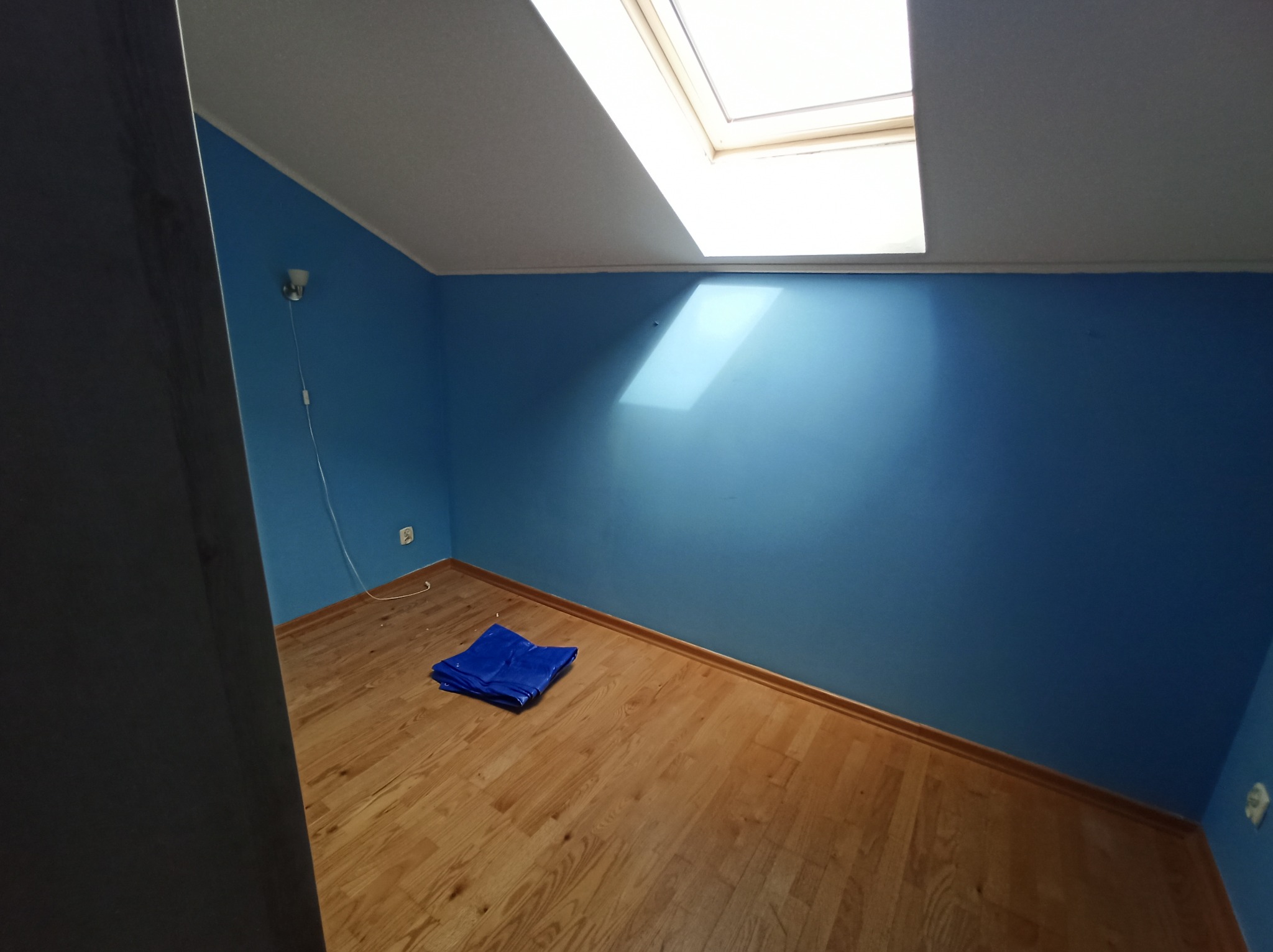 Pokój na poddaszu po malowaniu ścian na niebiesko, widoczne drewniane panele podłogowe i okno dachowe wpuszczające światło. Na podłodze leży złożona, niebieska folia malarska.