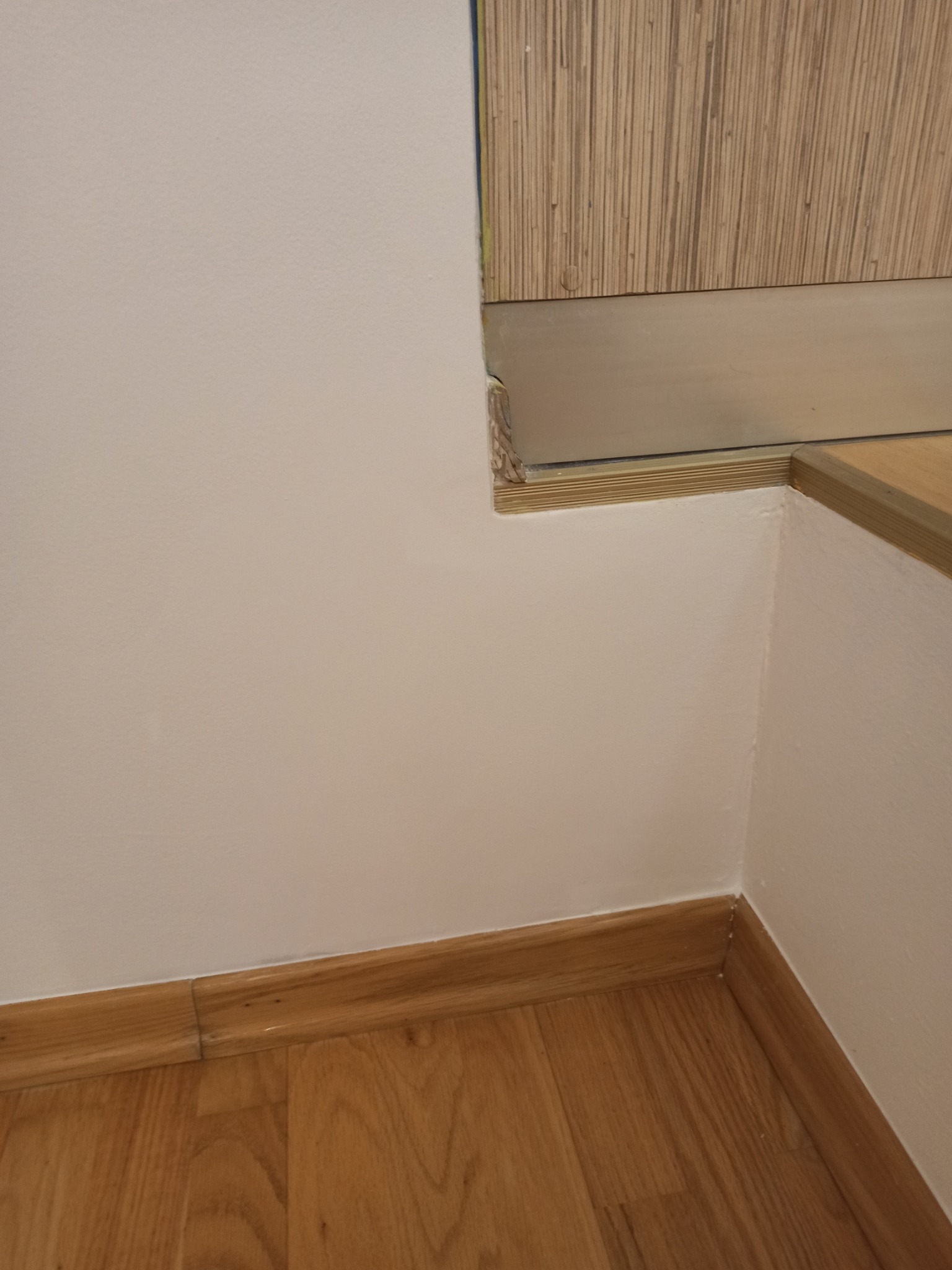 Ściana po malowaniu na biało, z drewnianą podłogą i listwami przypodłogowymi. Widoczny fragment schodów z drewnianym wykończeniem i tapetą na ścianie.