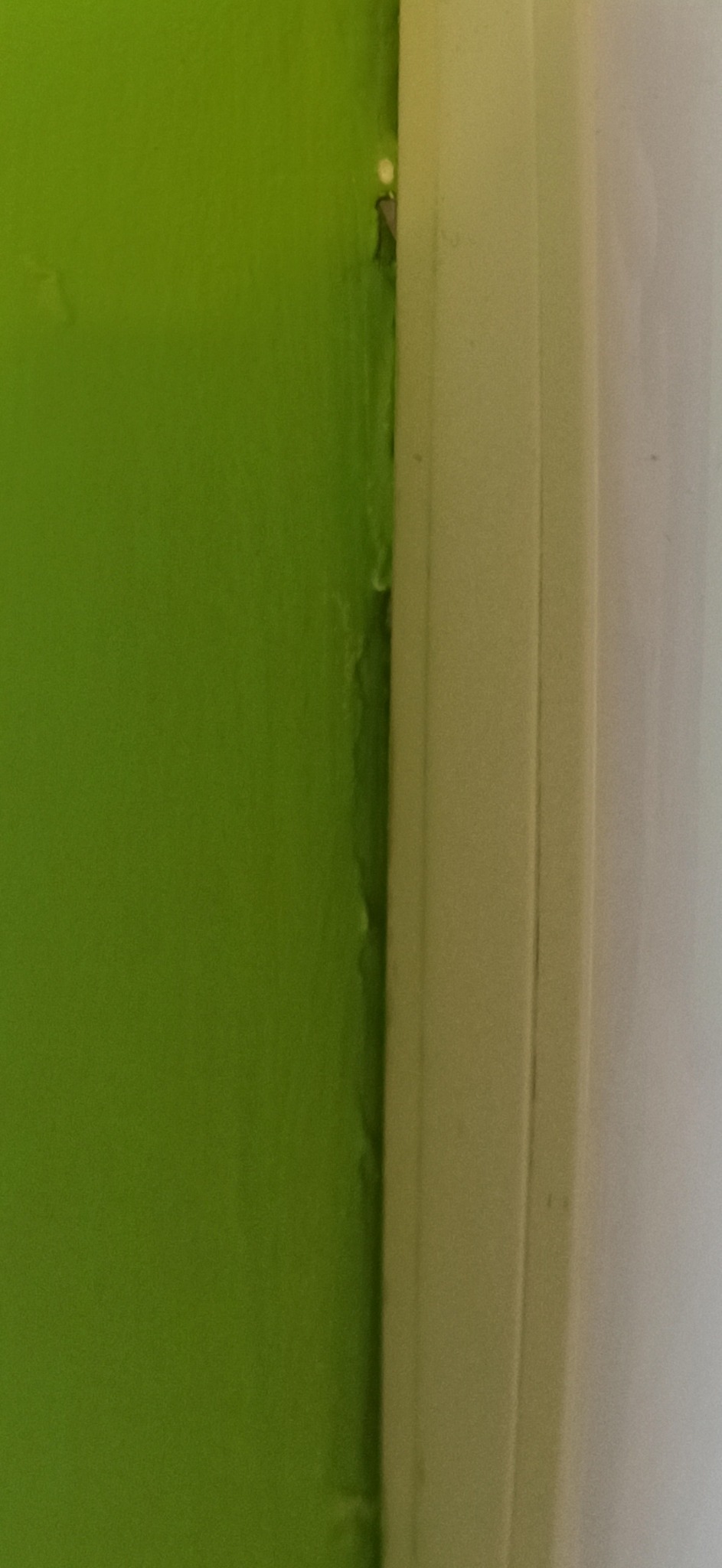 Fragment ściany w odcieniu zieleni, z widocznym niedokładnym wykończeniem przy listwie przypodłogowej. Widoczne nierówności i niedociągnięcia malarskie.