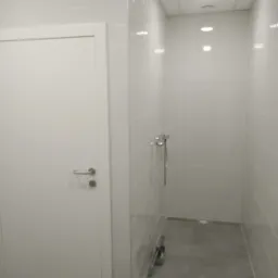 Nowoczesna, minimalistyczna łazienka z białymi płytkami i szarą podłogą, widoczna kabina prysznicowa z chromowaną armaturą oraz białe drzwi z metalową klamką.