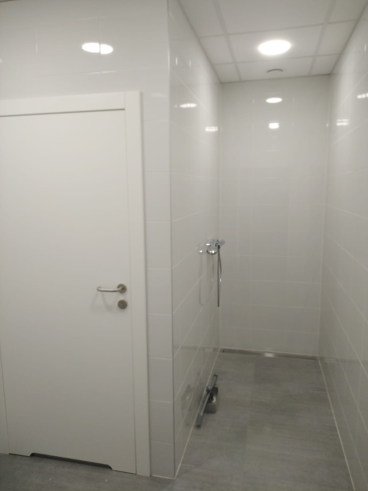 Nowoczesna, minimalistyczna łazienka z białymi płytkami i szarą podłogą, widoczna kabina prysznicowa z chromowaną armaturą oraz białe drzwi z metalową klamką.