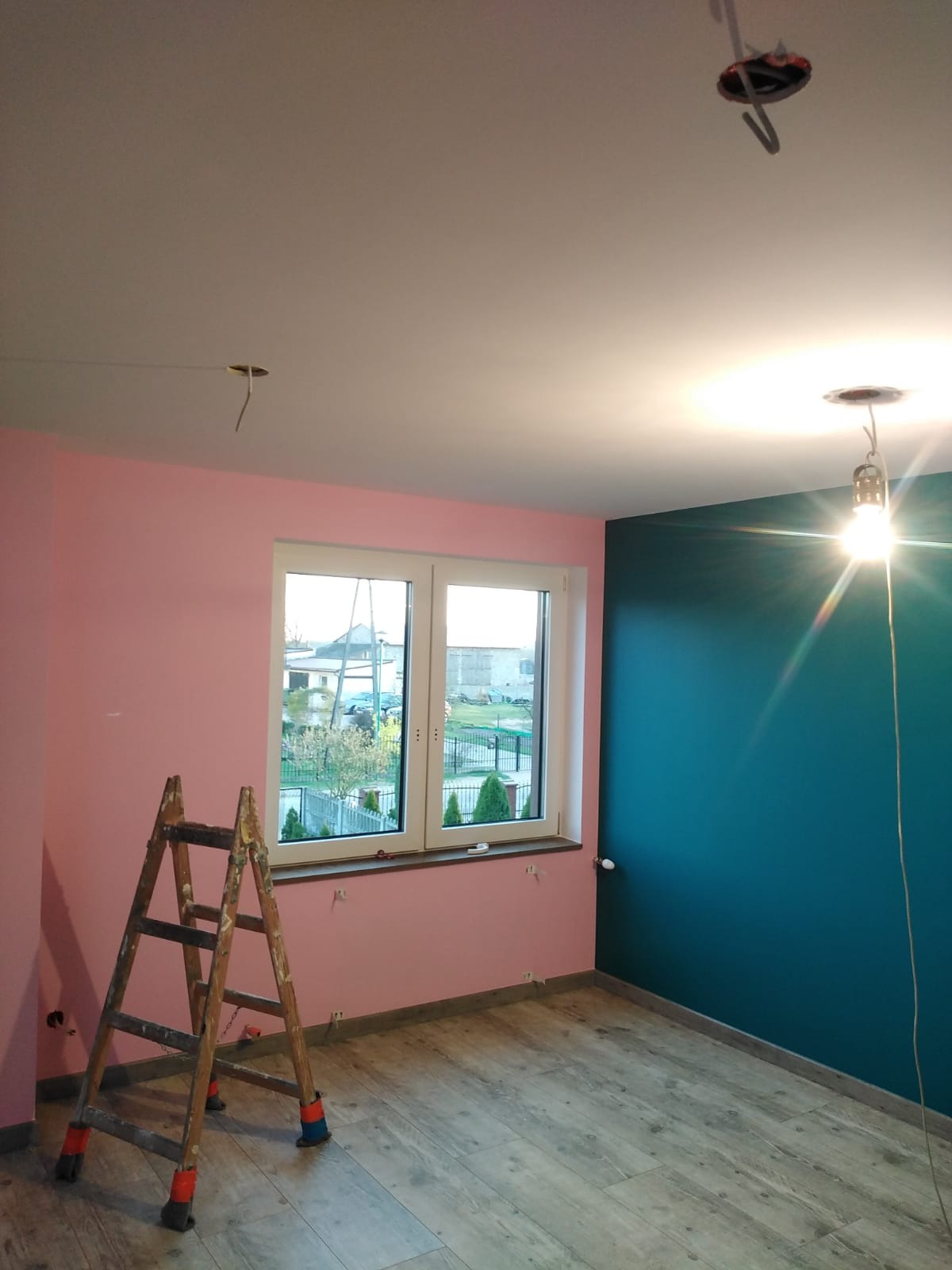 Pokój po malowaniu ścian na różowo i turkusowo, widoczna drewniana drabina, okno z widokiem na zewnątrz oraz wystające kable oświetleniowe z sufitu.