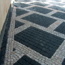 Nieregularny wzór z czarnej i szarej kostki granitowej ułożony w prostokątne segmenty, widok z góry, część kostki jeszcze nieukończona przy ścianie.