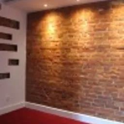 Wnętrze pokoju z ceglaną ścianą oświetloną punktowymi lampami sufitowymi, dekoracyjne wnęki w białej ścianie po lewej stronie, czerwona podłoga.