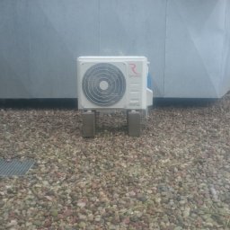 Klima-Rob Robert Majewski - Zewnętrzna jednostka klimatyzacji Rotenso Inverter, umieszczona na betonowych podstawach na dachu pokrytym drobnymi kamieniami, z widoczną kratką ściekową i fragmentem kartonu w tle.