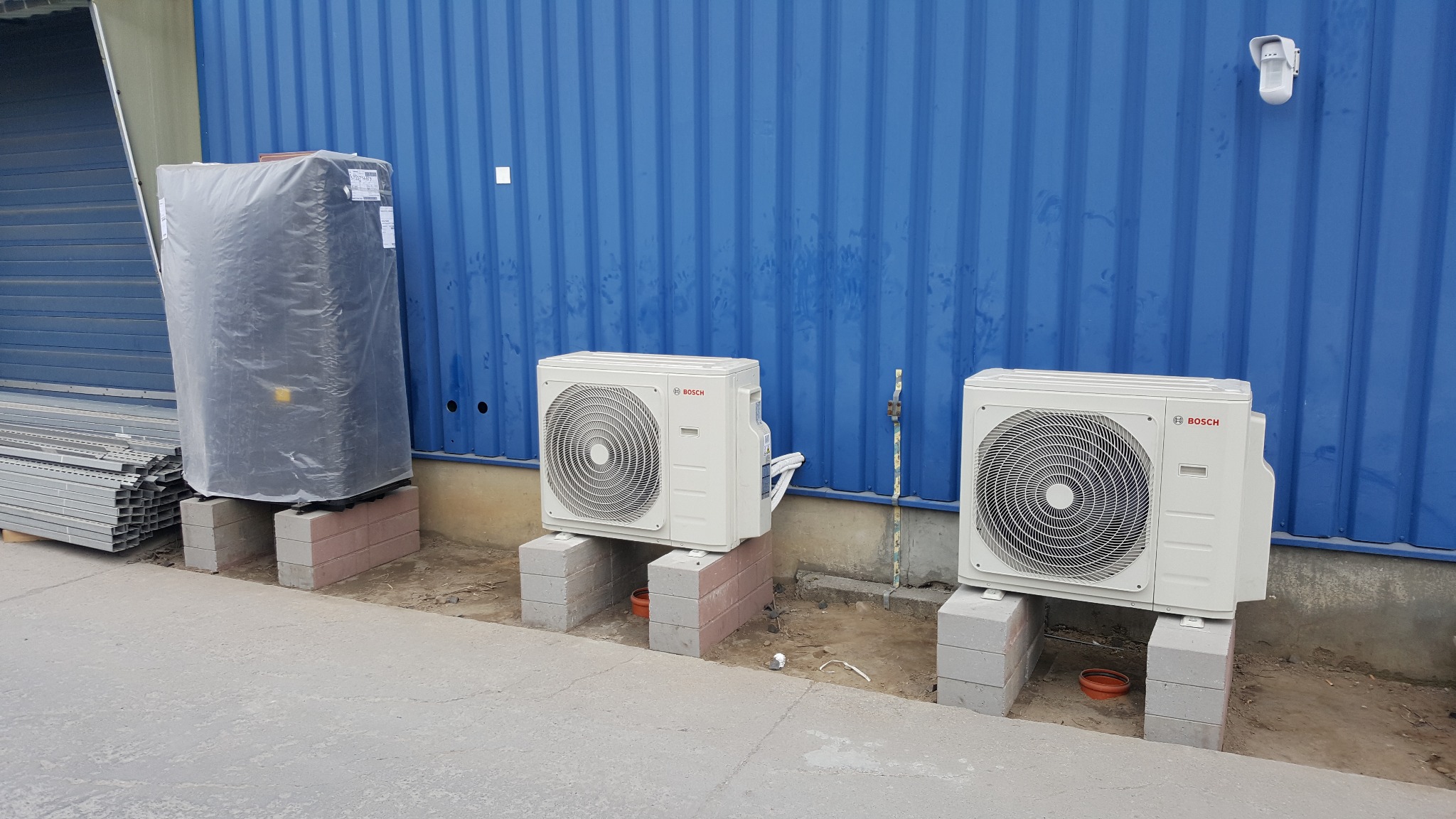 Dwa zewnętrzne agregaty klimatyzacji Bosch ustawione na bloczkach betonowych przed niebieską ścianą kontenera, obok duży zbiornik w folii ochronnej i stos metalowych profili.