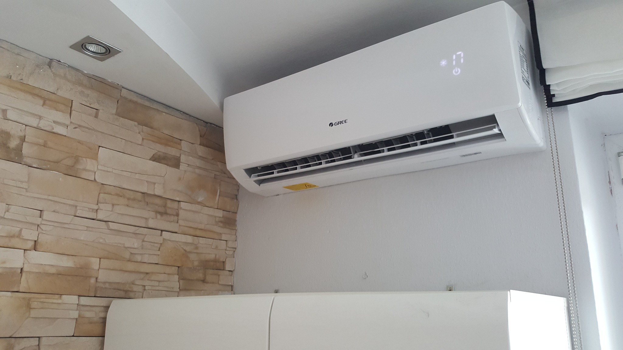 Biały klimatyzator marki Gree zamontowany na ścianie z dekoracyjną okładziną z piaskowca, z wyświetlaczem wskazującym temperaturę 17 stopni, widoczne białe meble pod spodem i roleta okienna z boku.