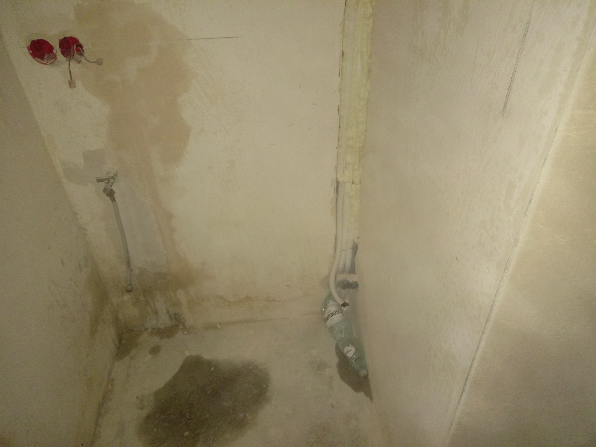 Wnętrze w trakcie remontu: surowe ściany z widocznymi śladami po instalacjach, odkryte kable elektryczne i rury zabezpieczone pianką montażową, butelka z wodą na podłodze.