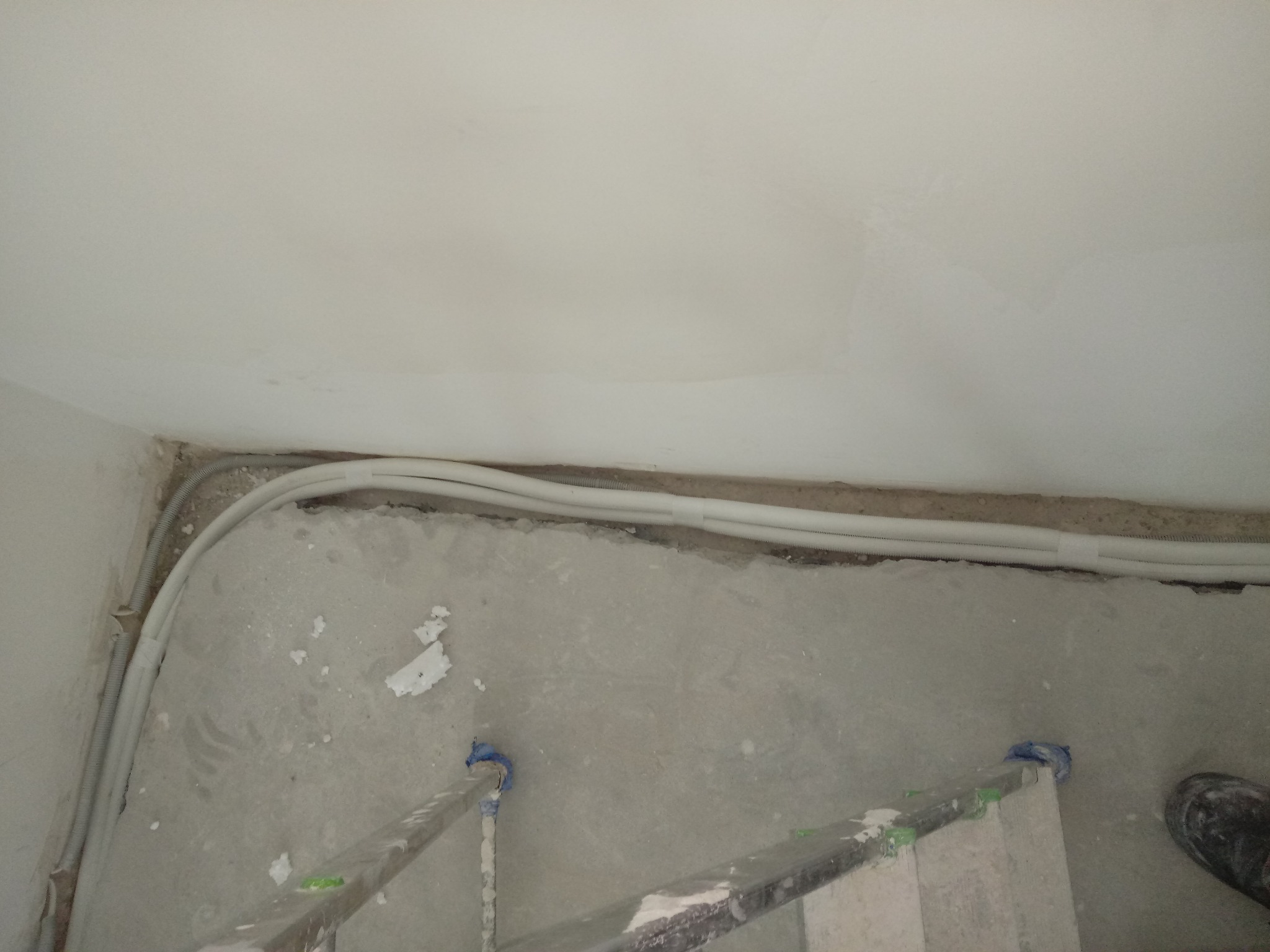 Białe rury instalacyjne prowadzone wzdłuż ściany i sufitu w trakcie remontu, widoczny fragment drabiny i buta roboczego.