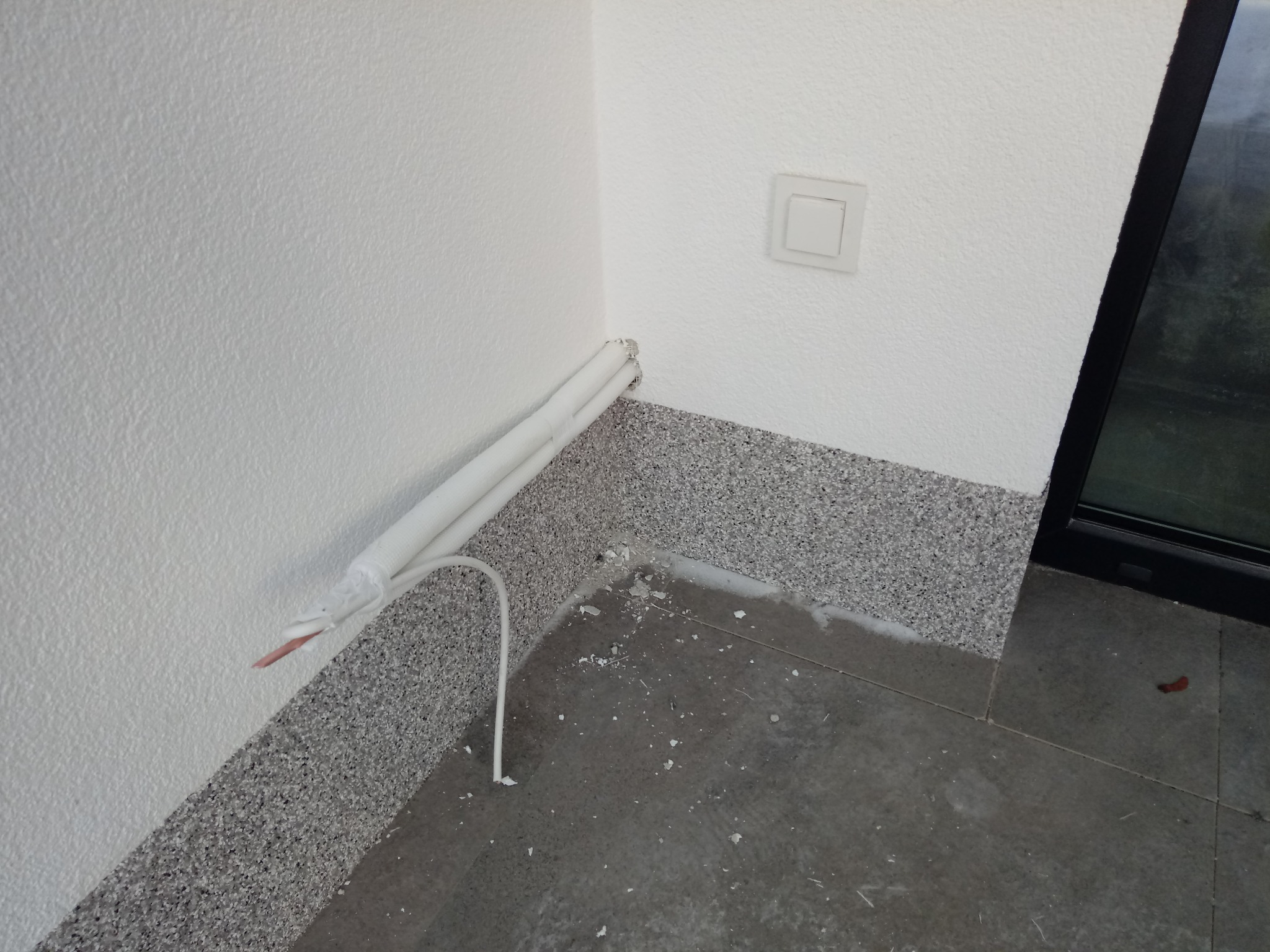 Wewnętrzny narożnik pomieszczenia z widocznymi rurami instalacji chłodniczej, częściowo zabezpieczonymi taśmą, wystające przewody, ślady wiercenia na podłodze i ścianie, szara podłoga z płytek...