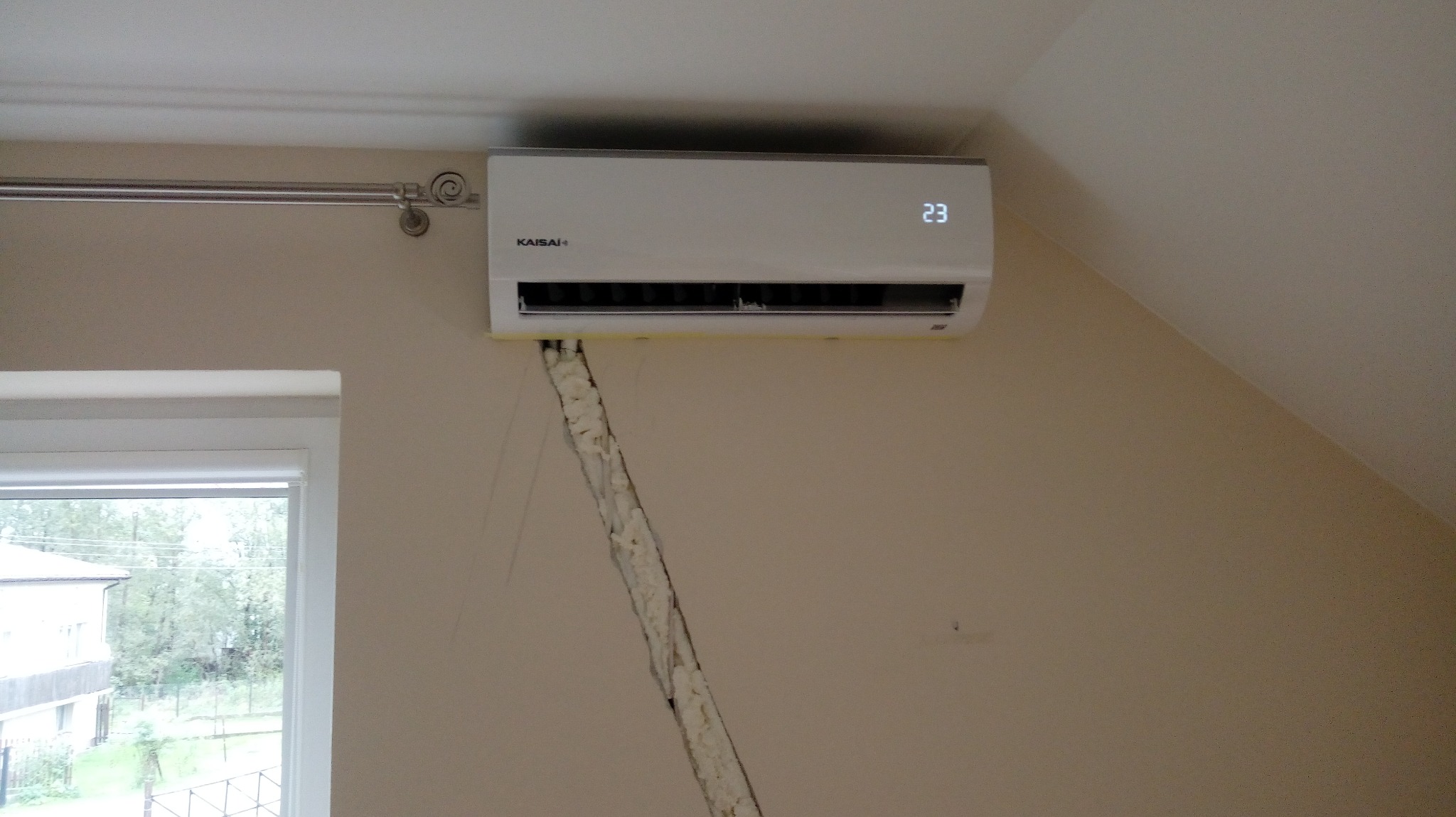 Biały klimatyzator Kaisai z wyświetlaczem temperatury 23 stopnie Celsjusza, zamontowany na beżowej ścianie w pokoju, z widoczną szczeliną wypełnioną pianką montażową, obok okna z widokiem na zewnątrz.