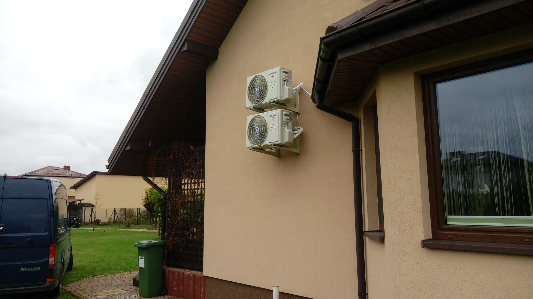 Dwa klimatyzatory marki Kaisai zamontowane jeden nad drugim na beżowej elewacji domu jednorodzinnego, widoczne rury odprowadzające skropliny, obok okno z brązową ramą, w tle niebieski samochód...