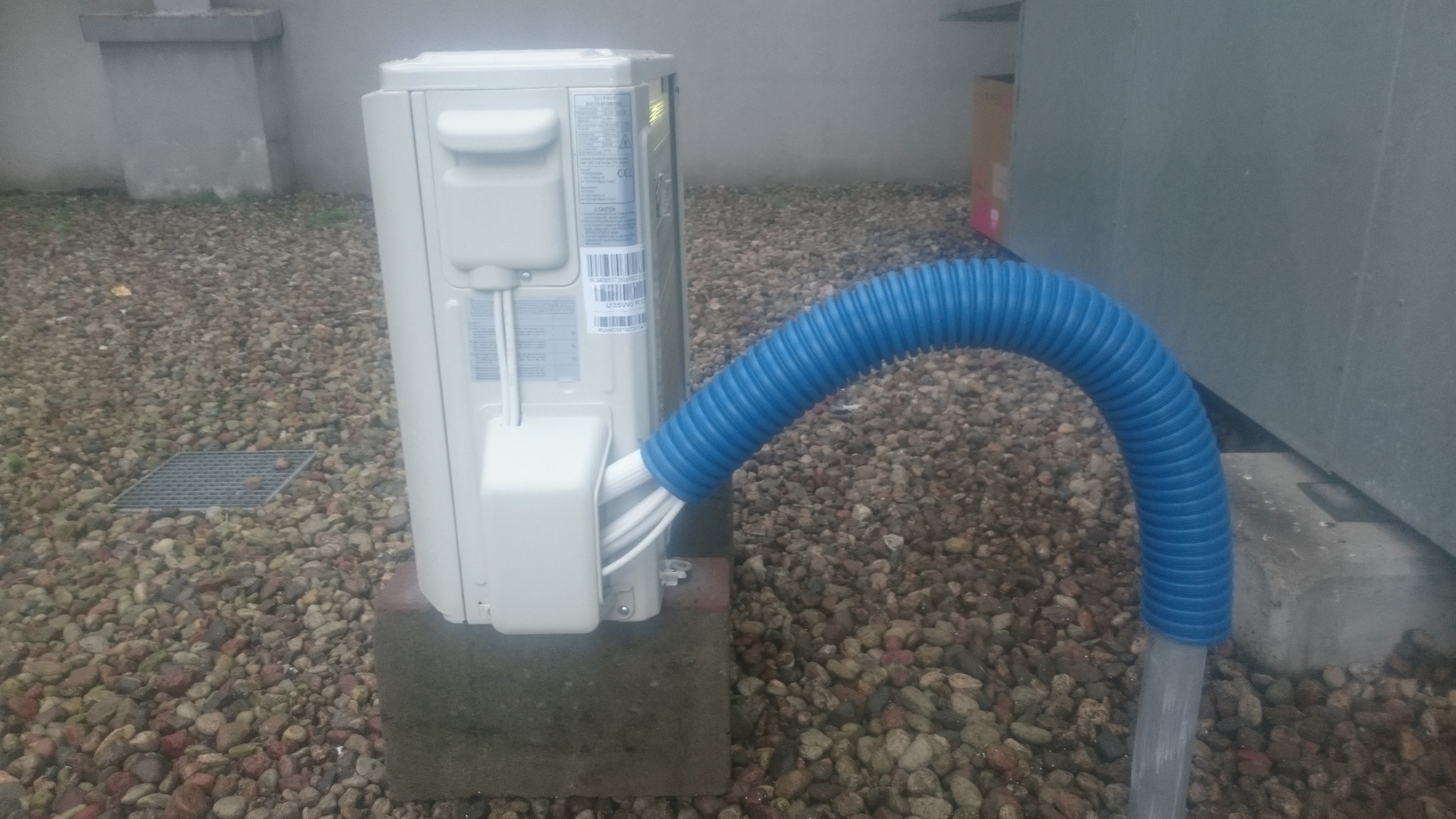 Zewnętrzna jednostka klimatyzacji na betonowym postumencie, widoczne podłączenie rur w niebieskiej karbowanej osłonie, tło z kamieni i betonowych elementów budynku.