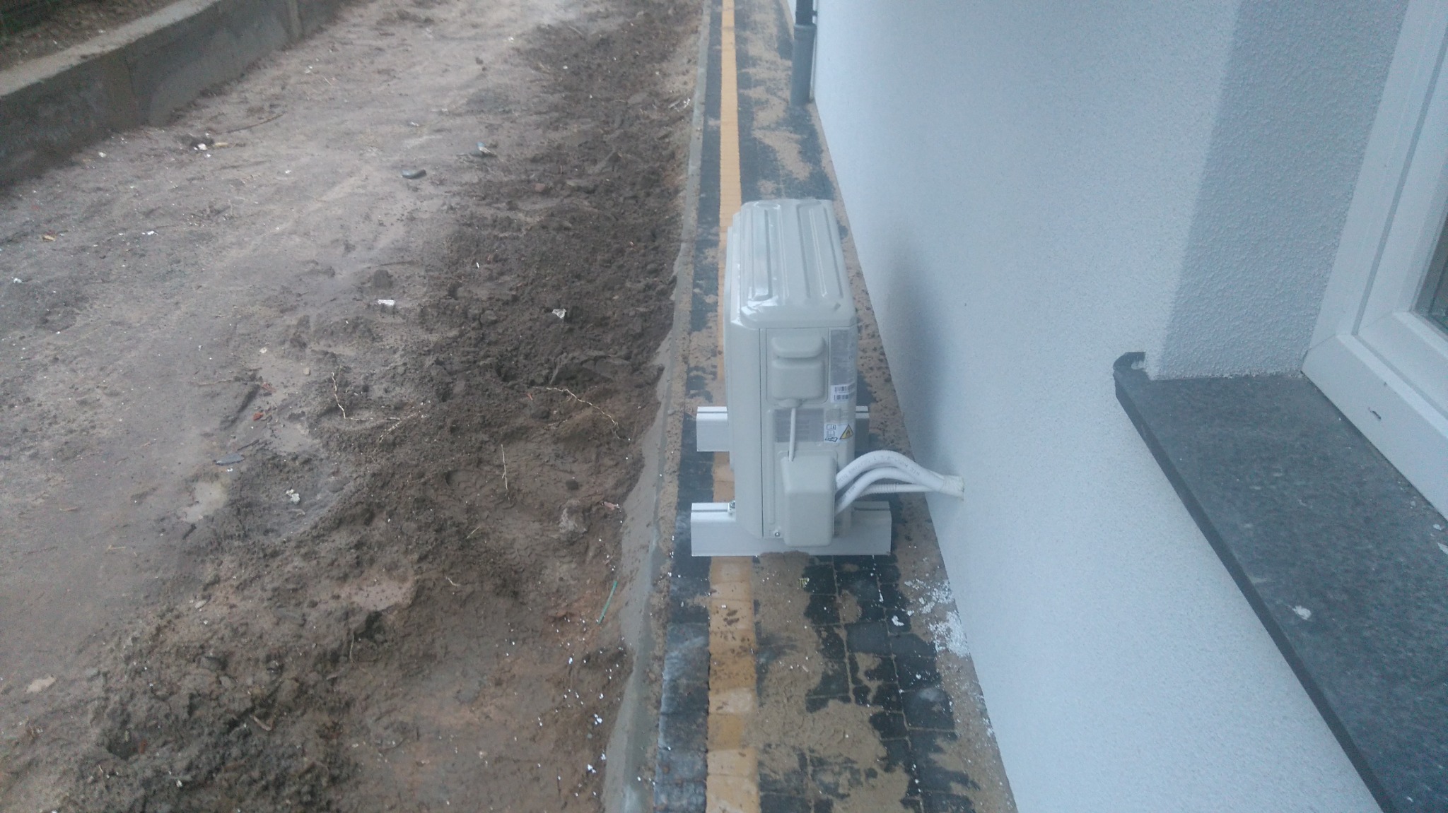 Zewnętrzna jednostka klimatyzacji zamontowana przy ścianie budynku, widoczne przewody, teren wokół w trakcie prac ziemnych.