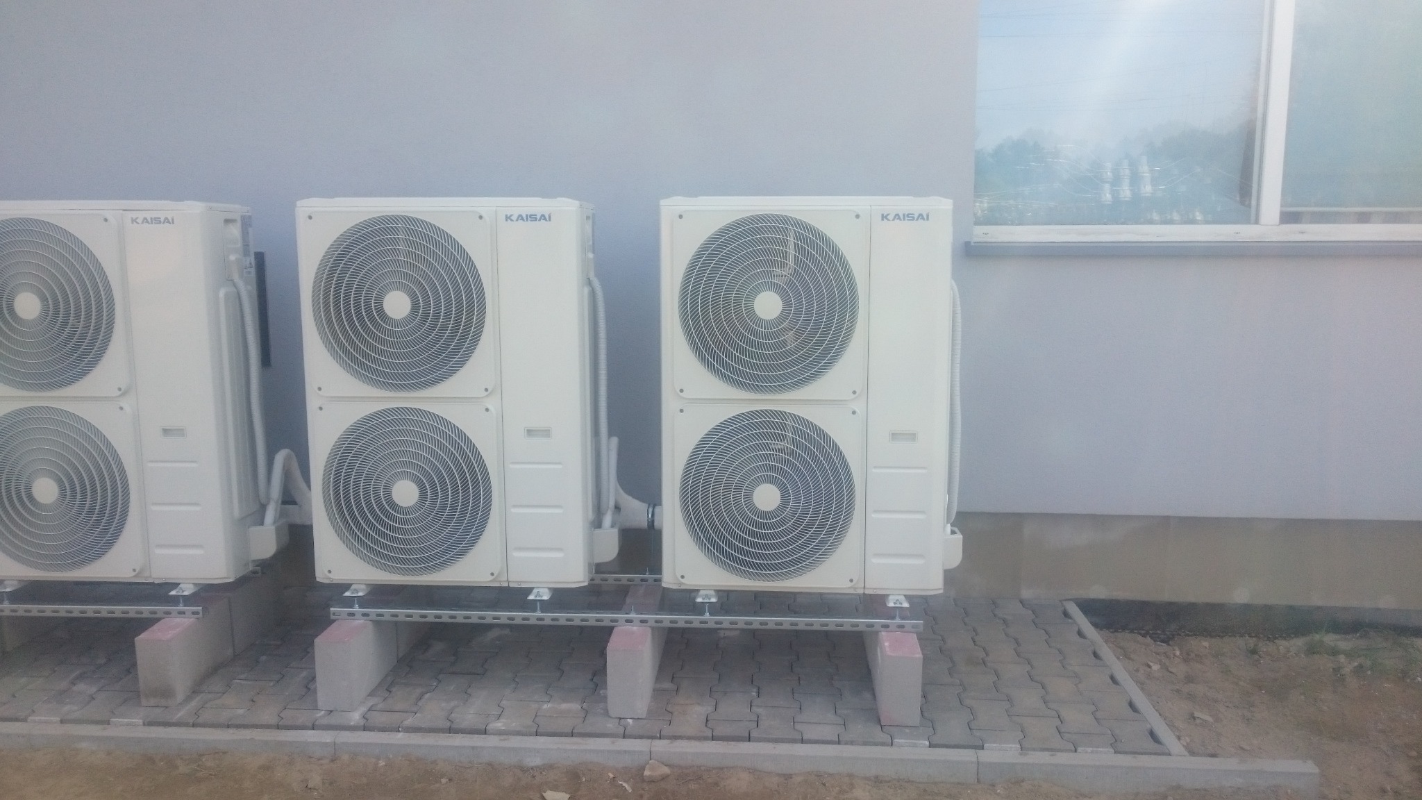 Zewnętrzne jednostki klimatyzacyjne Kaisai zamontowane na betonowych blokach i metalowych szynach przy szarej ścianie budynku z widocznym oknem.