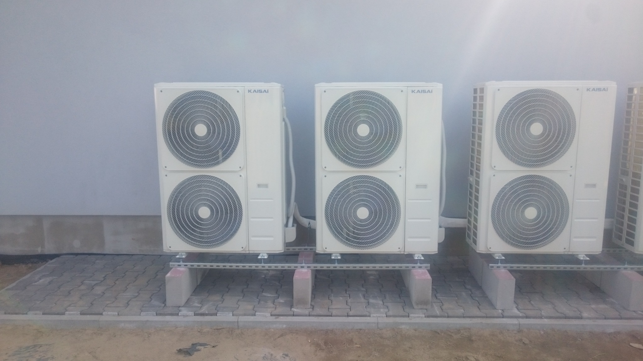 Trzy zewnętrzne jednostki klimatyzacyjne marki Kaisai ustawione na betonowych podstawach i metalowych szynach na wybrukowanym terenie przy szarej ścianie budynku.