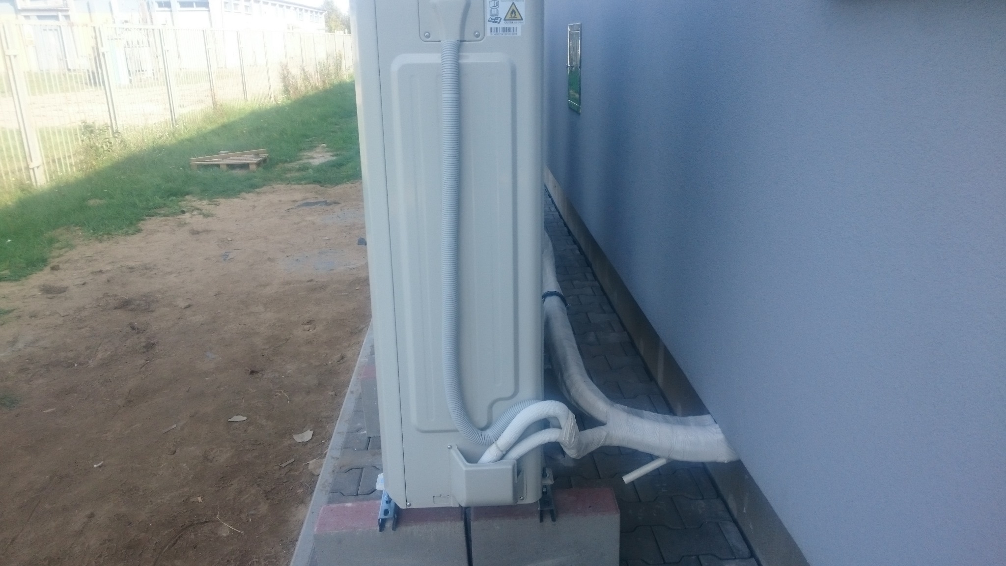 Zewnętrzna jednostka klimatyzacji zamontowana na betonowych bloczkach przy ścianie budynku, z widocznymi rurami odprowadzającymi skropliny i przewodem zasilającym, na tle trawnika i ogrodzenia.