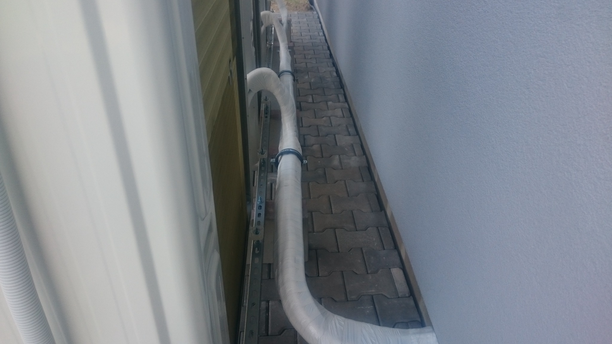 Zewnętrzna jednostka klimatyzacji z izolowanymi rurami chłodniczymi, zamocowanymi do ściany budynku, widoczne kostki brukowe na chodniku.