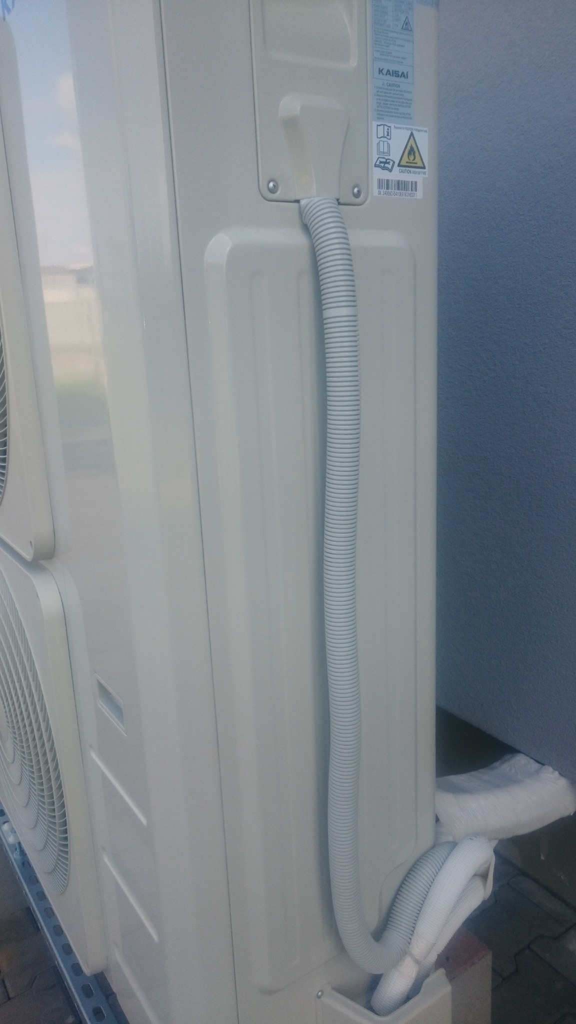 Zewnętrzna jednostka klimatyzacji Kaisai z widocznymi przewodami odprowadzającymi skropliny, zamontowana przy szarej ścianie budynku.