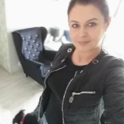 Kobieta w czarnej kurtce pozuje w salonie z ciemnym fotelem z guzikami i małym czarnym psem z różową obrożą.