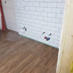 Łazienka w trakcie wykańczania: ułożone drewnopodobne płytki podłogowe z krzyżykami dystansowymi, białe kafelki na ścianie z wyprowadzonymi instalacjami wodno-kanalizacyjnymi, widoczna poziomica...