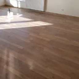 Nowo ułożona podłoga z paneli w odcieniach brązu, widoczne promienie słoneczne wpadające przez okno, grzejnik pod oknem, puszka po farbie przy ścianie.