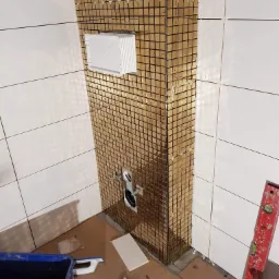 Fragment łazienki w trakcie wykańczania z zamontowaną instalacją podtynkową WC obłożoną złotą mozaiką, widoczne krzyżyki dystansowe, poziomica i pojemnik z narzędziami.