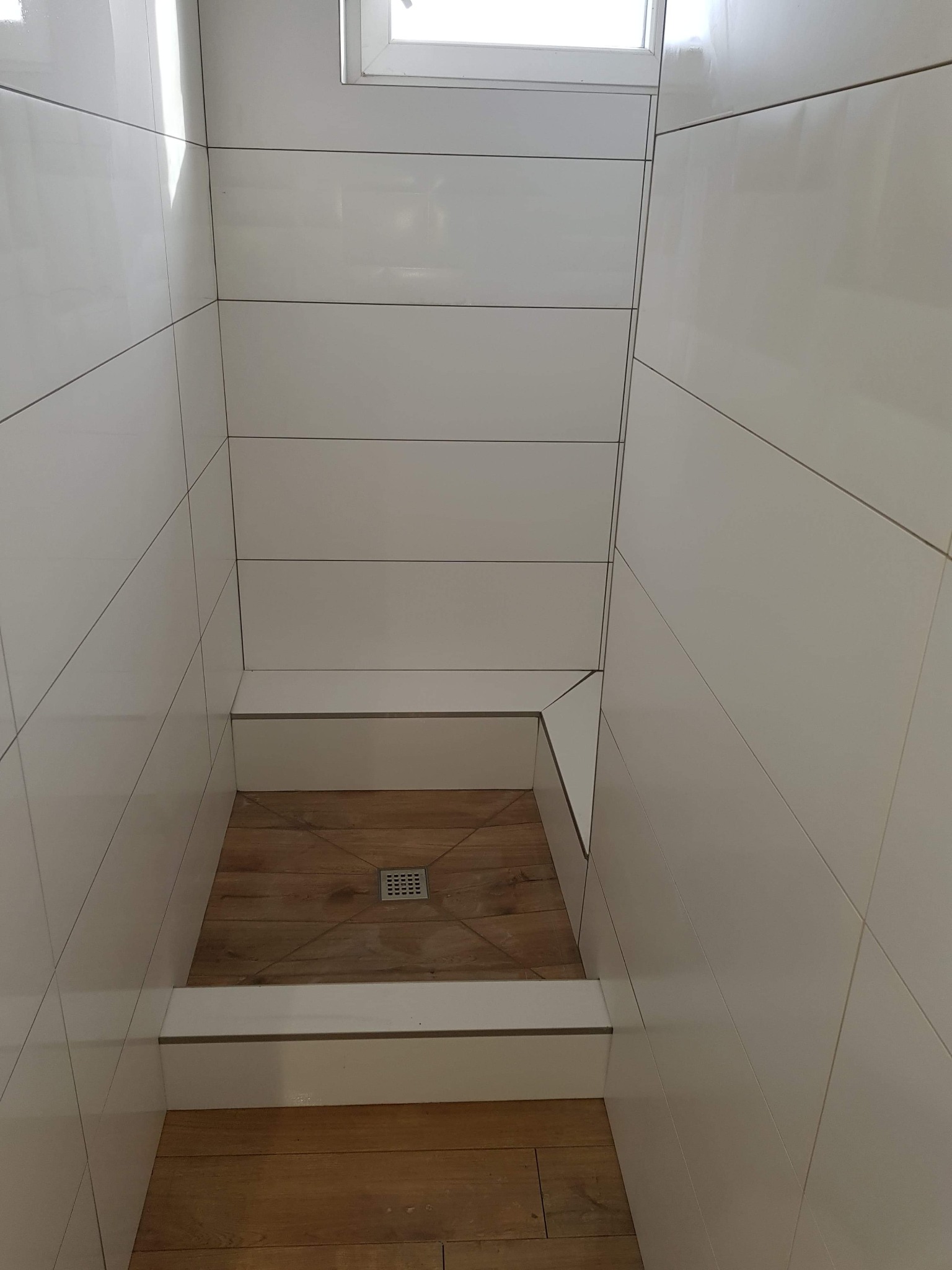 Nowoczesna kabina prysznicowa walk-in z białymi, prostokątnymi płytkami ceramicznymi ułożonymi poziomo na ścianach i drewnopodobną podłogą z odpływem liniowym w centralnej części.