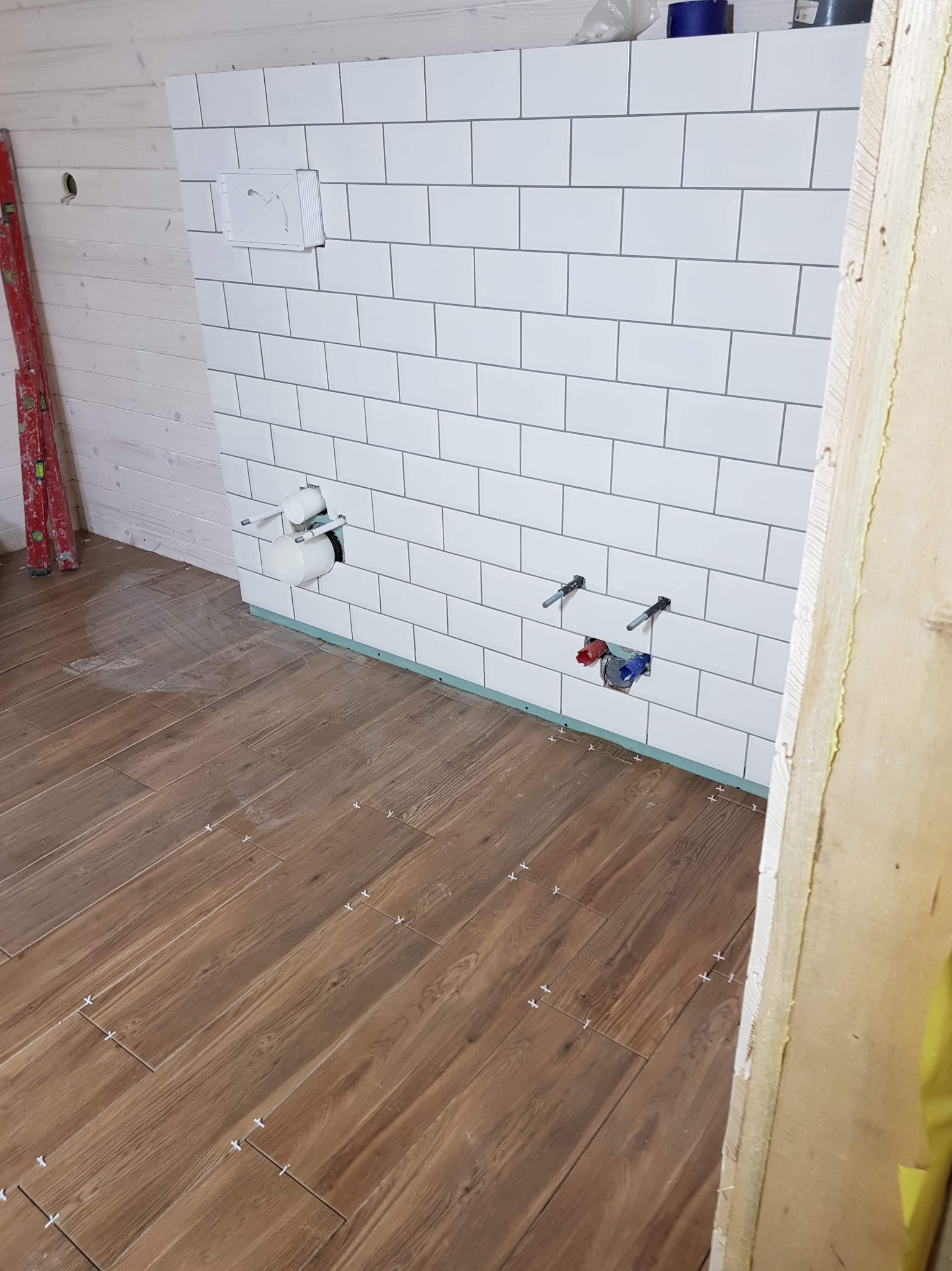 Łazienka w trakcie wykańczania: ułożone drewnopodobne płytki podłogowe z krzyżykami dystansowymi, białe kafelki na ścianie z wyprowadzonymi instalacjami wodno-kanalizacyjnymi, widoczna poziomica...