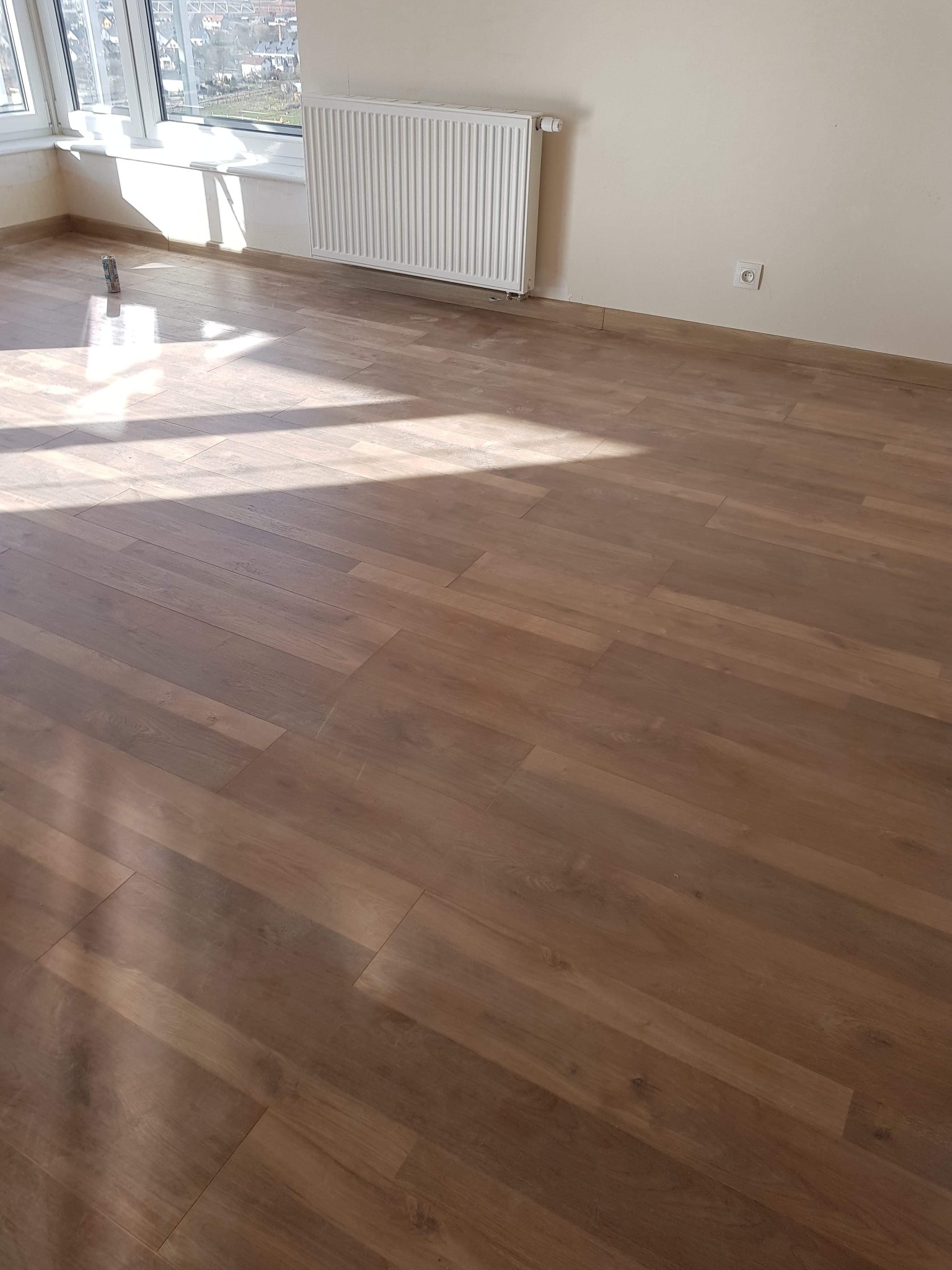 Nowo ułożona podłoga z paneli w odcieniach brązu, widoczne promienie słoneczne wpadające przez okno, grzejnik pod oknem, puszka po farbie przy ścianie.