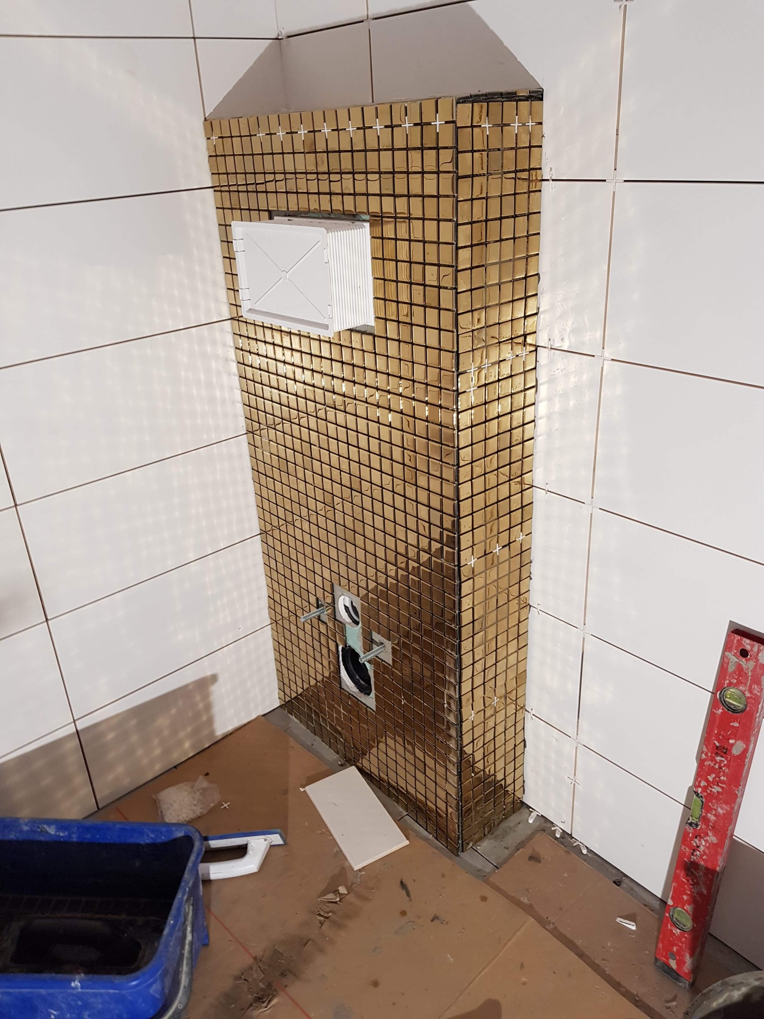 Fragment łazienki w trakcie wykańczania z zamontowaną instalacją podtynkową WC obłożoną złotą mozaiką, widoczne krzyżyki dystansowe, poziomica i pojemnik z narzędziami.