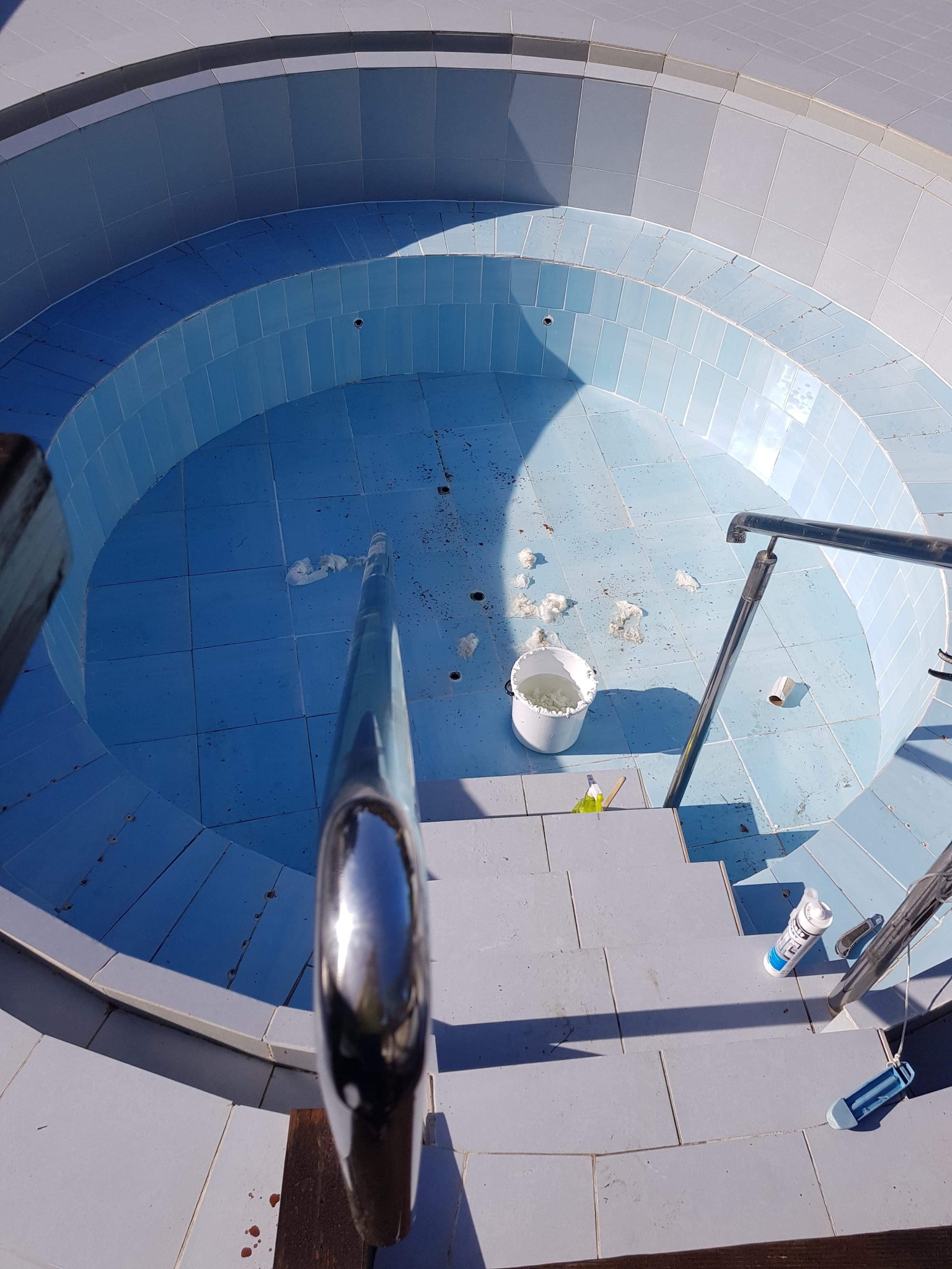 Pusty, okrągły basen wyłożony błękitnymi płytkami, z widocznymi schodkami i metalową barierką, w trakcie czyszczenia, z białym wiadrem i porozrzucanymi kawałkami materiału wewnątrz.