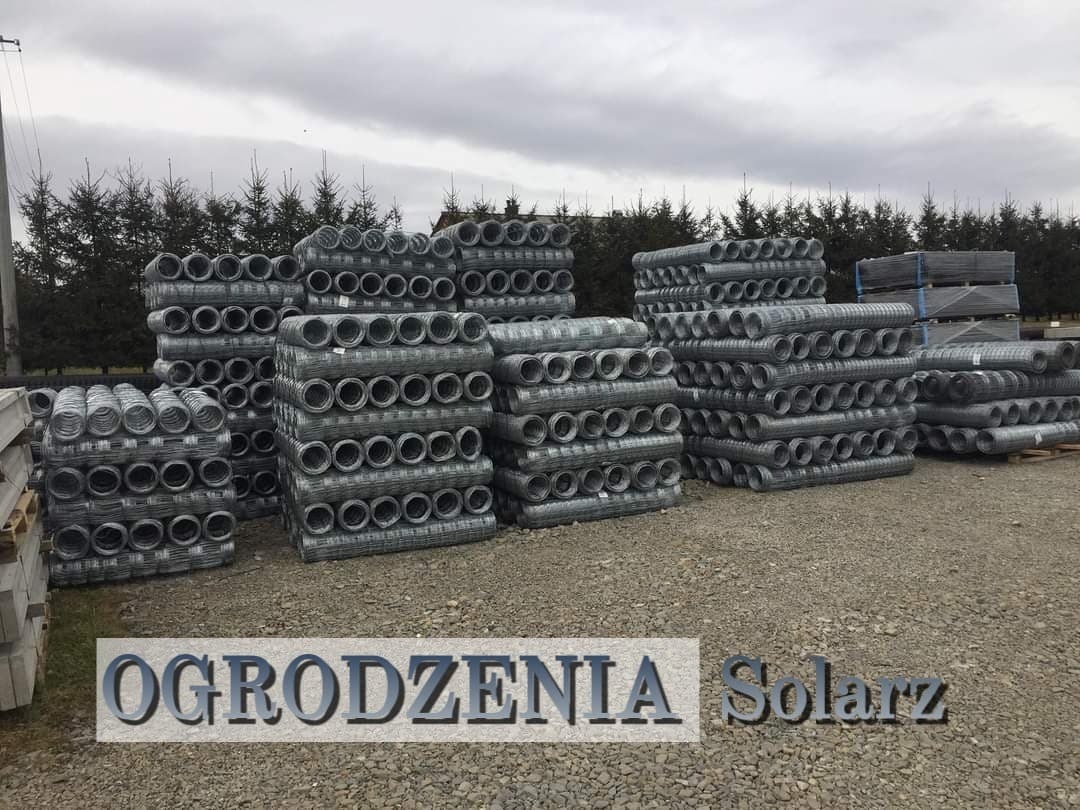 Stosy zwiniętej siatki ogrodzeniowej, przygotowane do transportu na żwirowym placu, widoczne betonowe słupki i iglaki w tle, pochmurne niebo.