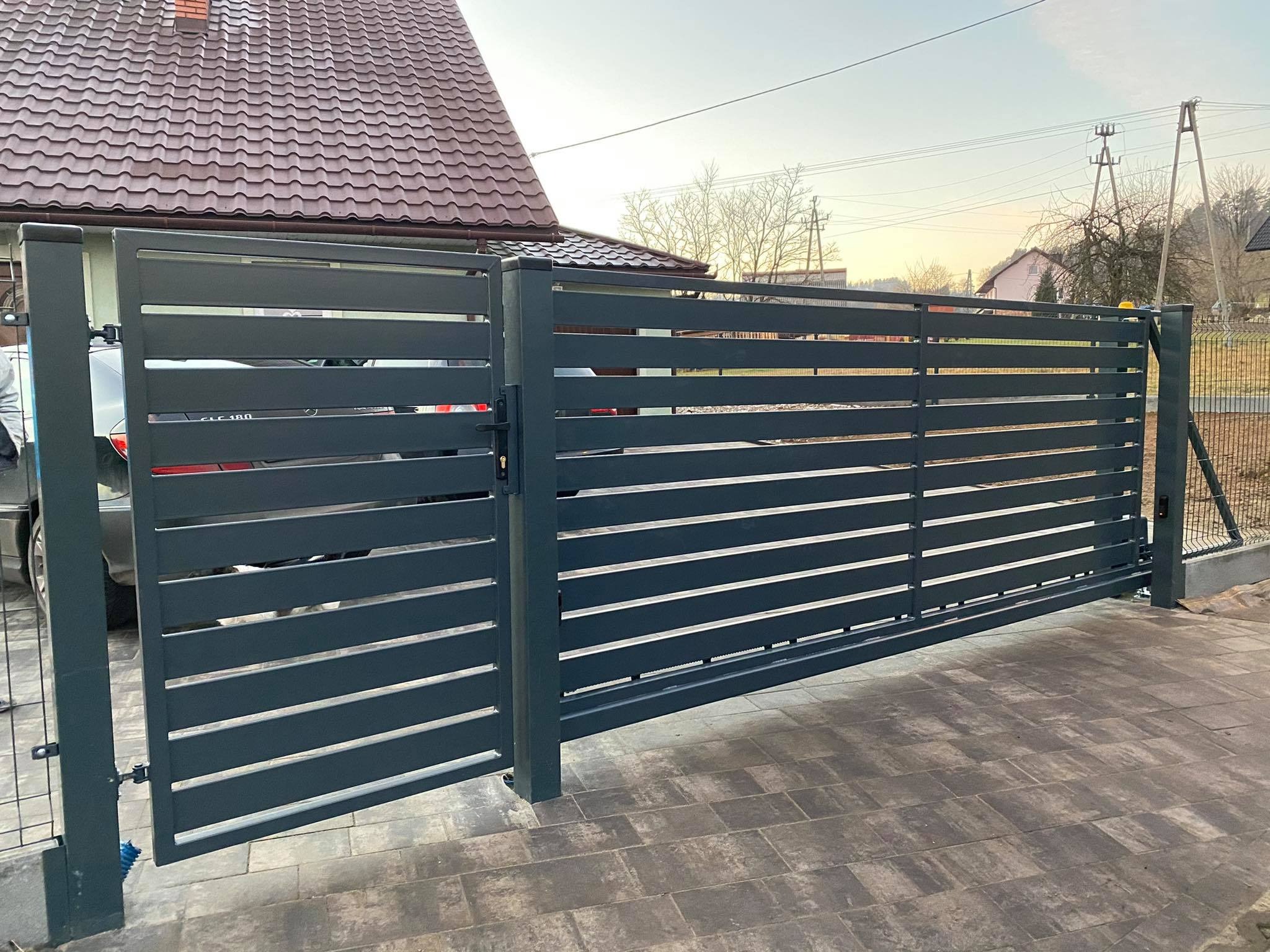 Nowoczesna, przesuwna brama w kolorze antracytowym z poziomymi panelami, częściowo otwarta, z widocznym samochodem i domem w tle.