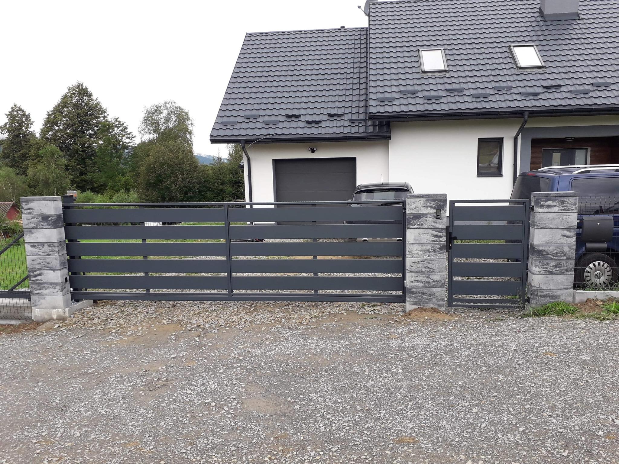 Nowoczesna, przesuwna brama w kolorze antracytowym z poziomymi panelami, osadzona na betonowych słupkach z szarego kamienia łupanego, na tle białego domu z ciemnoszarą dachówką i szarym garażem.