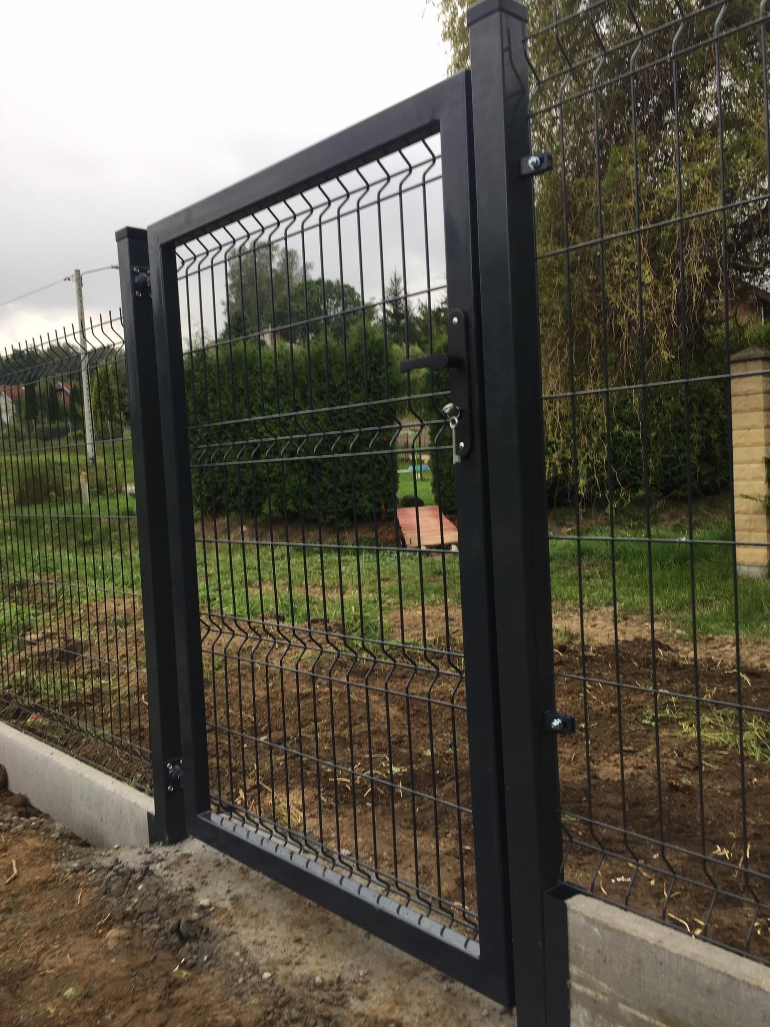 Nowo zamontowana, czarna, metalowa brama panelowa z zamkiem i klamką, widoczna na tle zielonego ogrodu i nieba z chmurami.