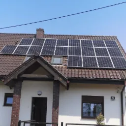 Dom jednorodzinny z brązowym dachem i panelami słonecznymi, widok zza czarnego ogrodzenia z numerem domu 29 po prawej stronie.