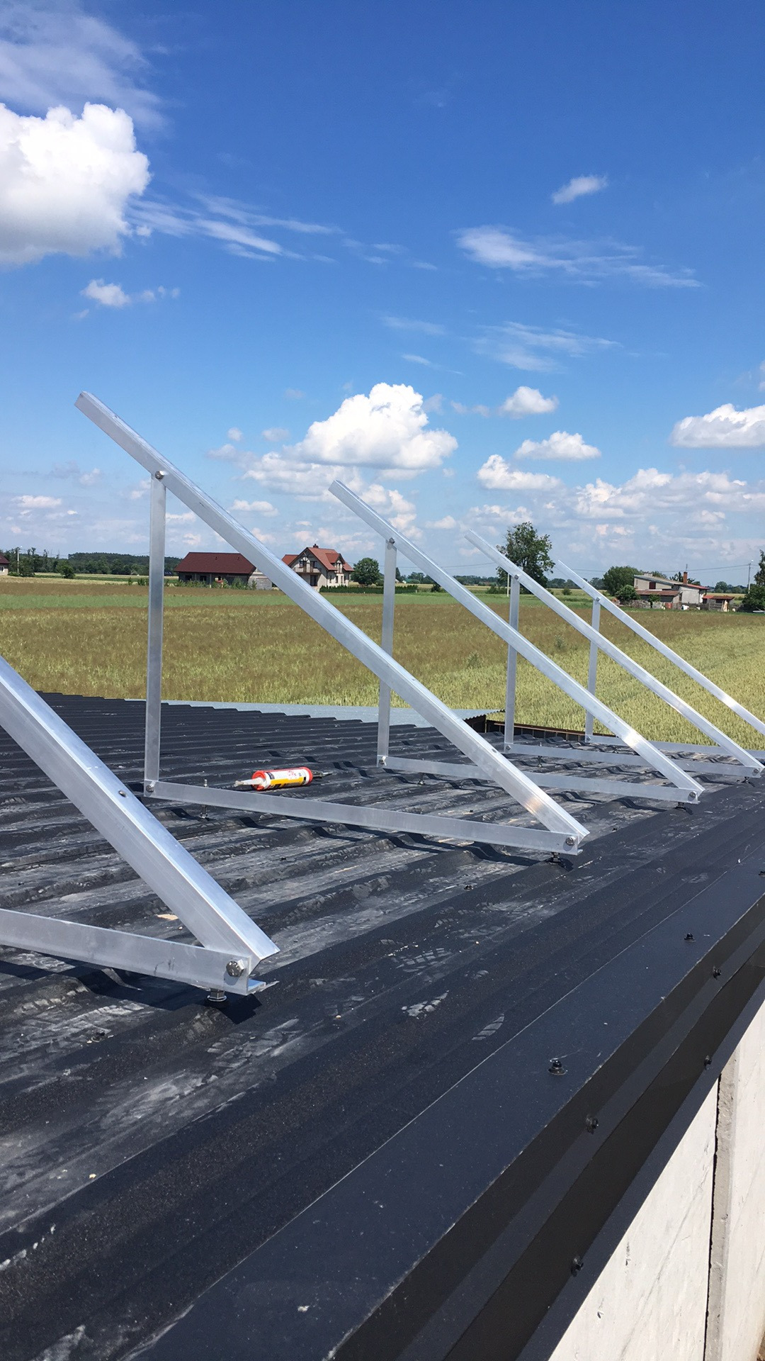 Montaż konstrukcji wsporczej pod panele słoneczne na dachu z blachy trapezowej, widoczna tuba kleju montażowego, w tle krajobraz wiejski z domami.