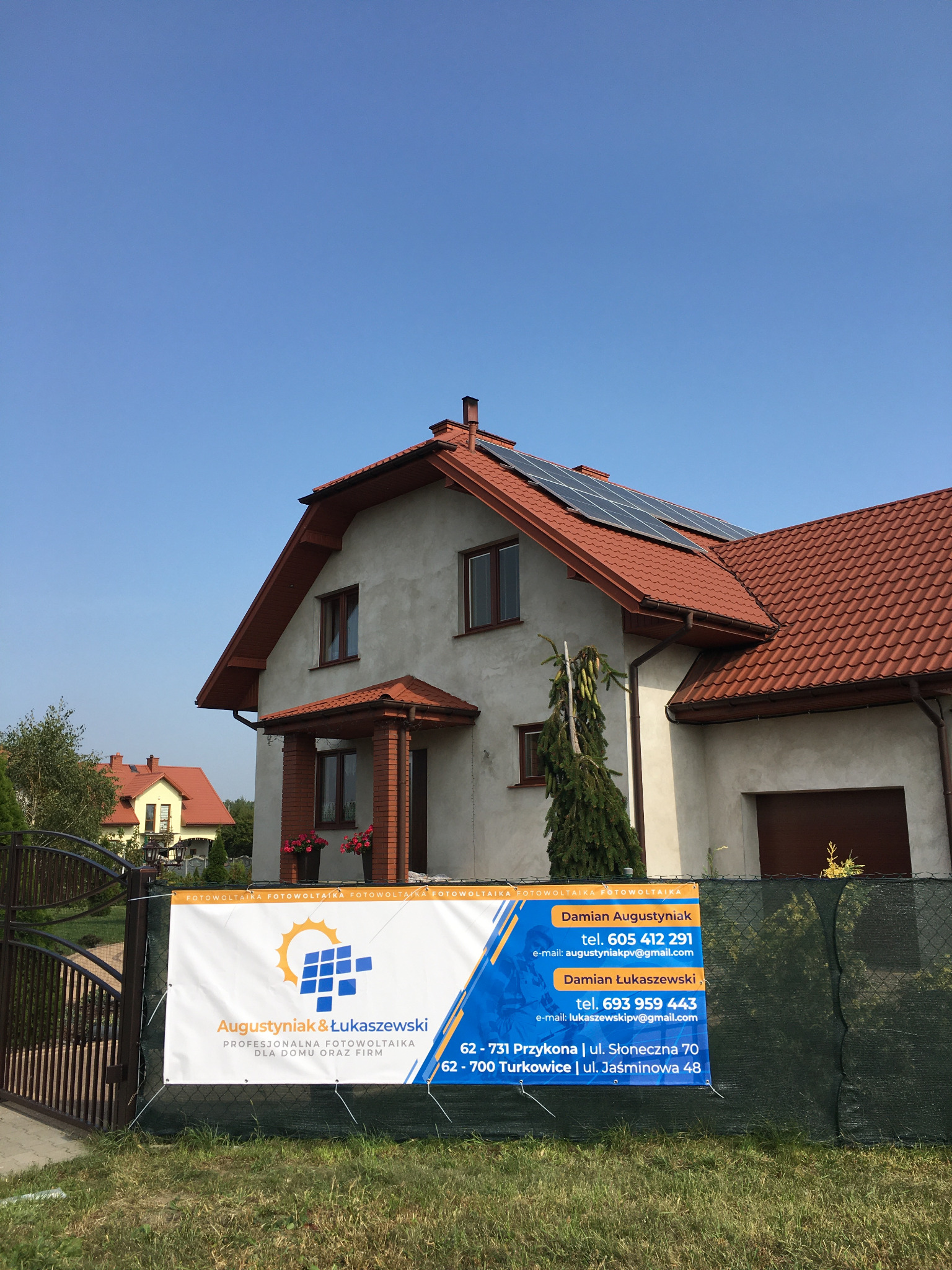 Dom jednorodzinny z panelami słonecznymi na dachu, widoczny baner reklamowy firmy fotowoltaicznej na ogrodzeniu.