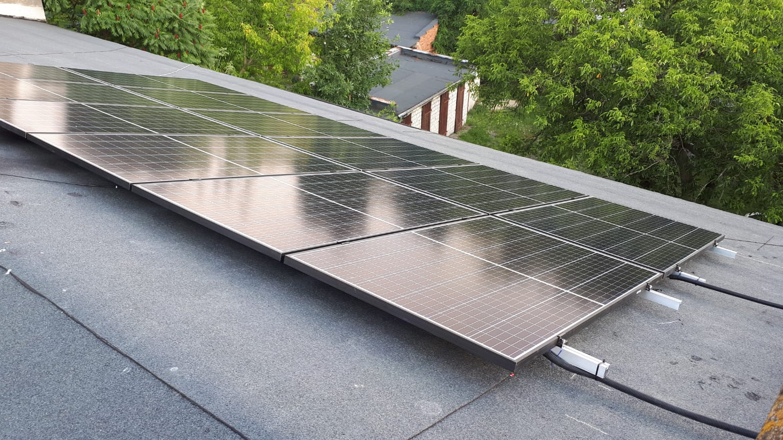 Panele słoneczne zamontowane na płaskim dachu pokrytym papą, widoczne kable i elementy mocujące, w tle zieleń drzew.