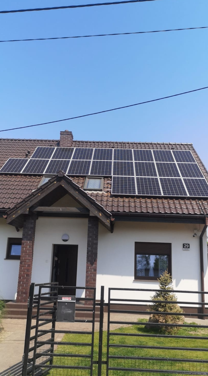 Dom jednorodzinny z brązowym dachem i panelami słonecznymi, widok zza czarnego ogrodzenia z numerem domu 29 po prawej stronie.