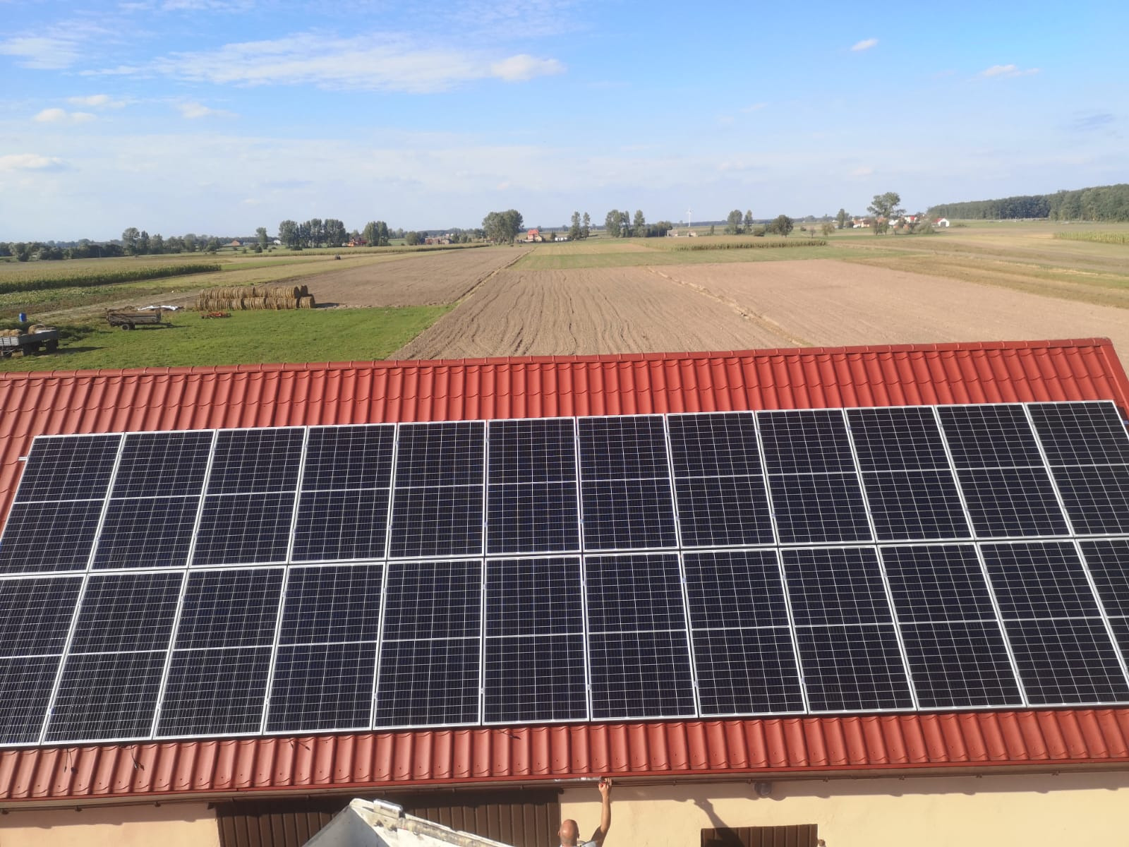 Panel fotowoltaiczny na czerwonym dachu domu jednorodzinnego, widok z góry na wiejski krajobraz z polami i belami siana.