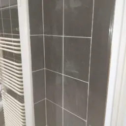 Fragment ściany wyłożonej ciemnymi, błyszczącymi płytkami ceramicznymi z jasną fugą, obok biały grzejnik drabinkowy i fragment białej ościeżnicy drzwiowej.